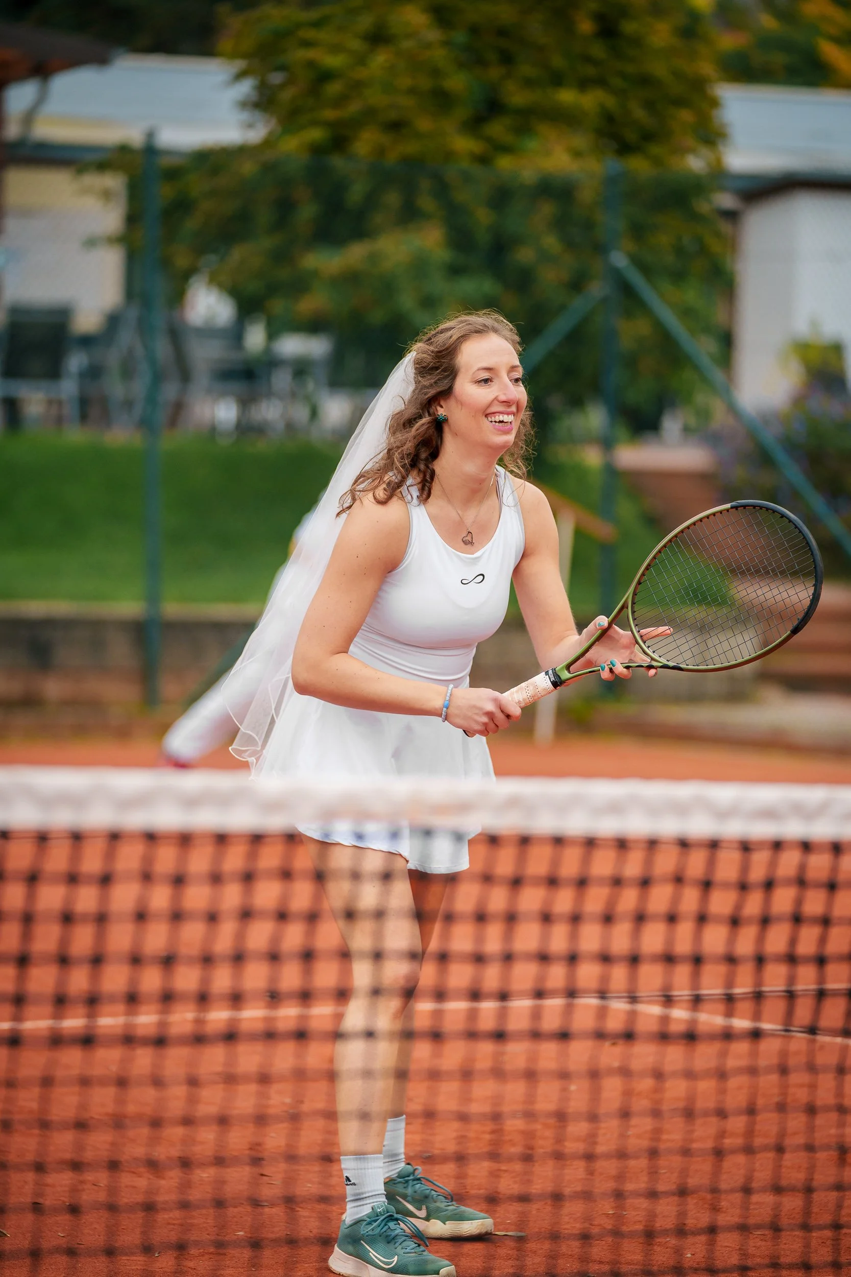 Eine Frau in einem weißen Kleid und Hornbrille spielt Tennis auf einem roten Tennisplatz, trägt einen Schleier und lächelt.