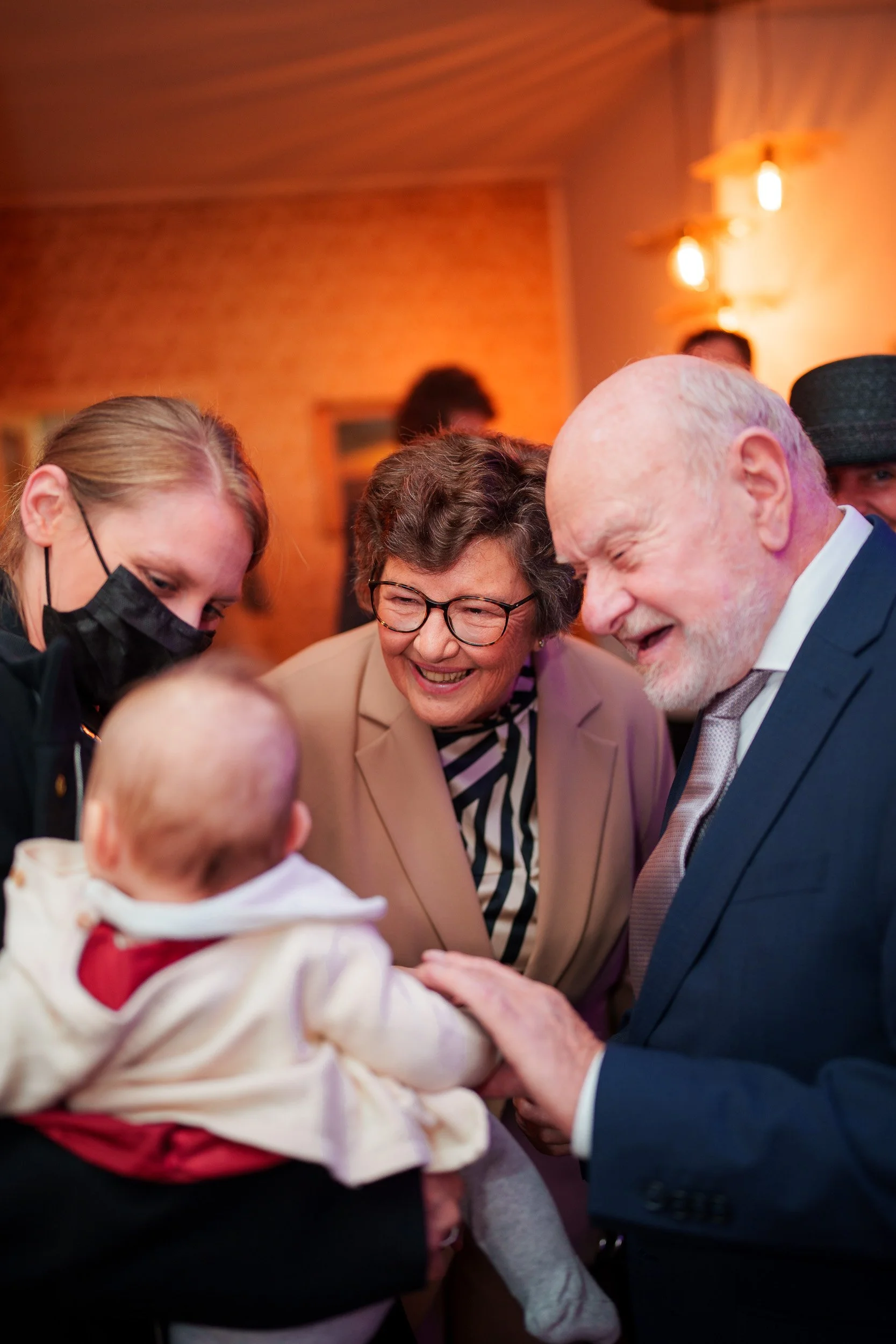 Eine Gruppe von vier Menschen, zwei ältere Erwachsene, eine jüngere Frau mit Maske, lächeln und schauen auf ein Baby, das von einer Person gehalten wird, in einem warm beleuchteten Raum.