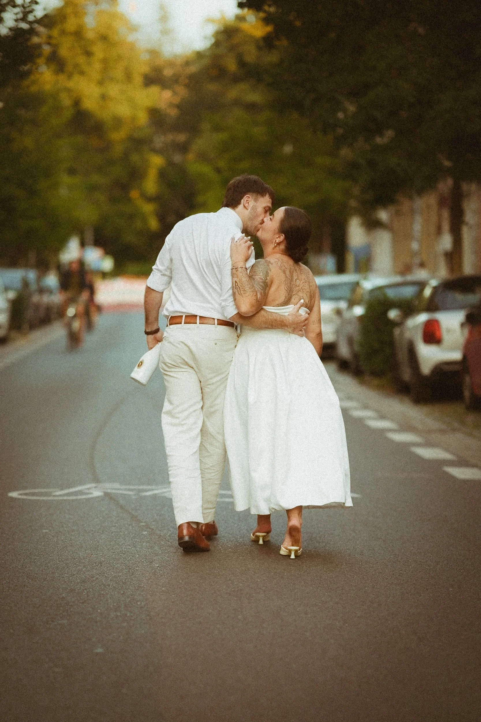 Ein Paar küsst sich auf einer Straße in einem weißen Kleidung, die Frau hat Tattoos auf ihren Armen und Rücken, im Hintergrund sind grüne Bäume und parkende Autos.