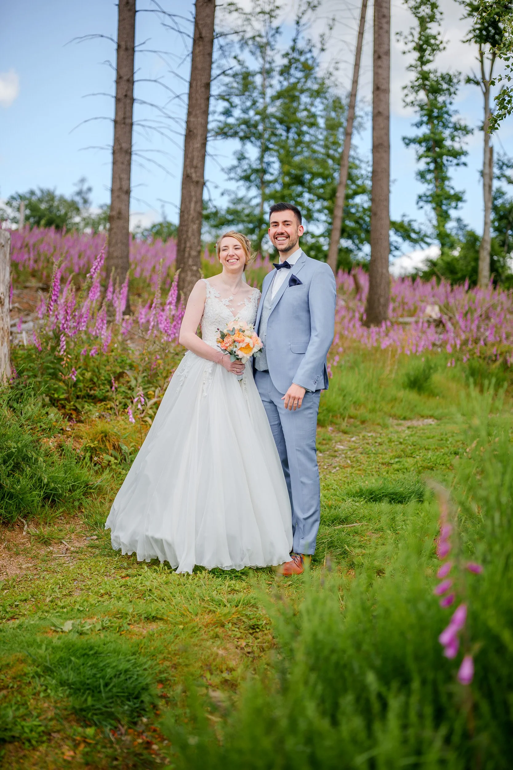 Ein glückliches Brautpaar steht im Grünen mit pinken Blumen im Hintergrund.