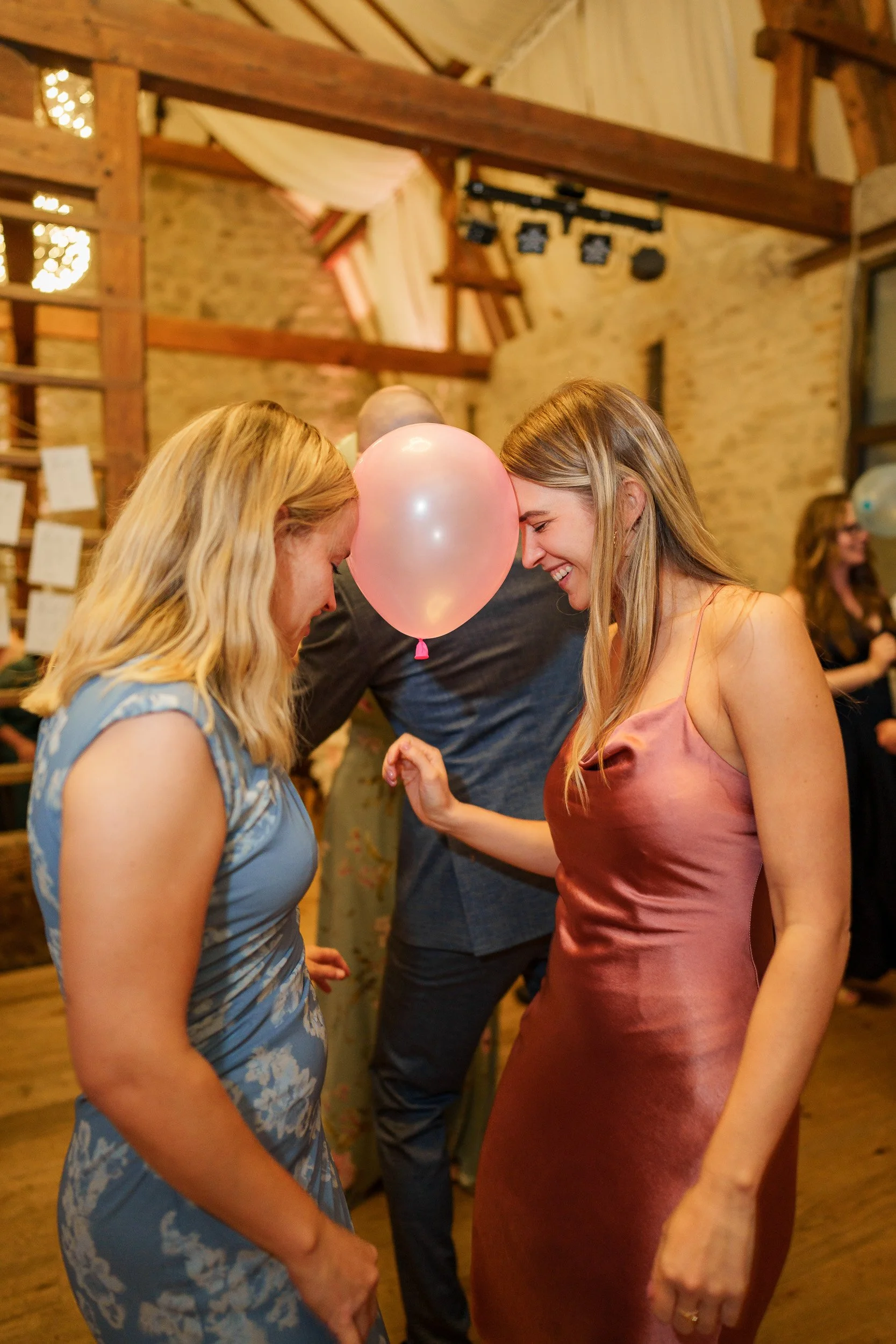 Zwei lachende Frauen bei einer Party, die mit einem Ballon in der Mitte beide Köpfe berühren, in einem rustikalen Raum mit Holzstruktur.