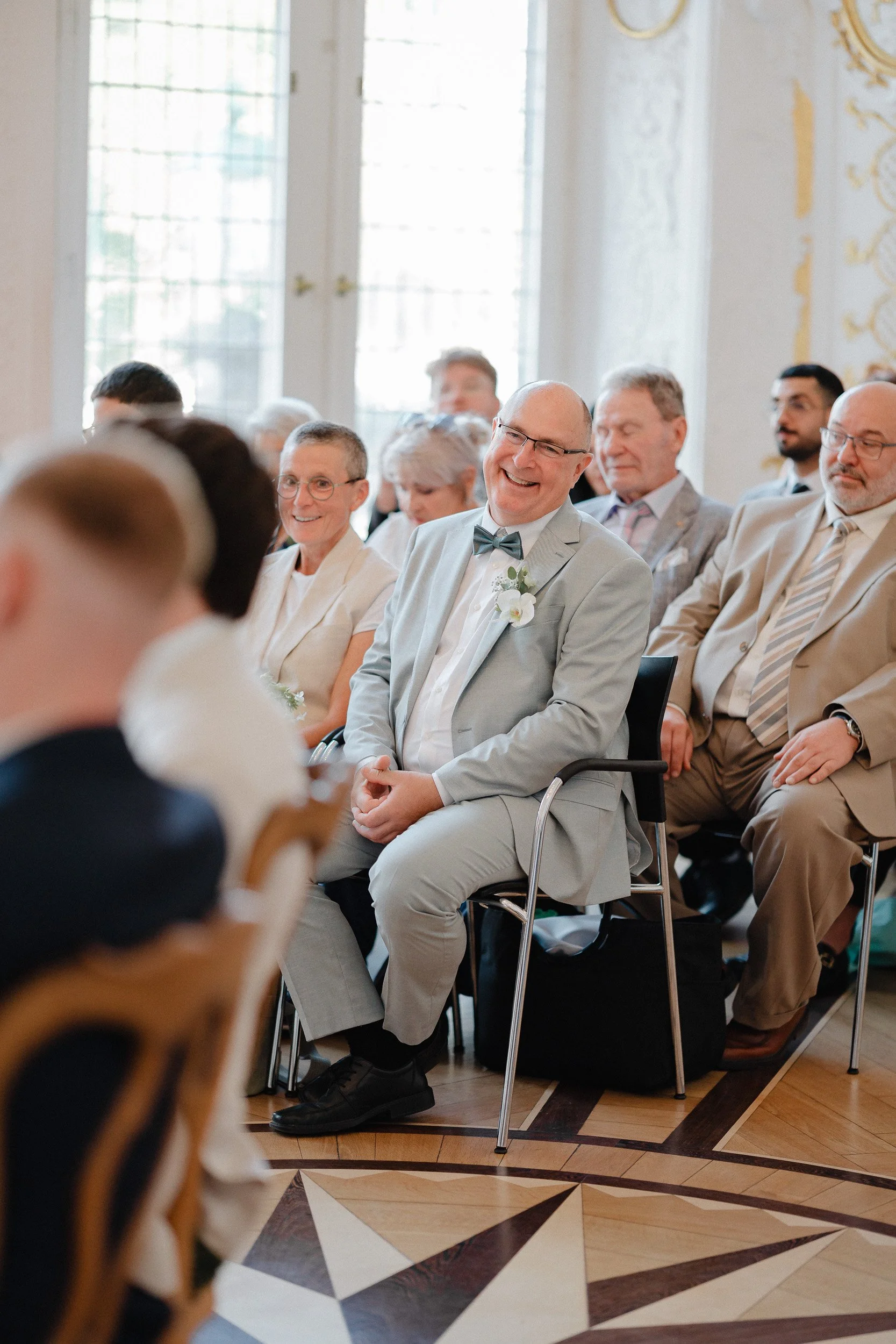 Hochzeitsszene mit lachendem Bräutigam in hellgrauem Anzug und Schleife, sitzend in einem eleganten Raum, umgeben von Gästen in formeller Kleidung.