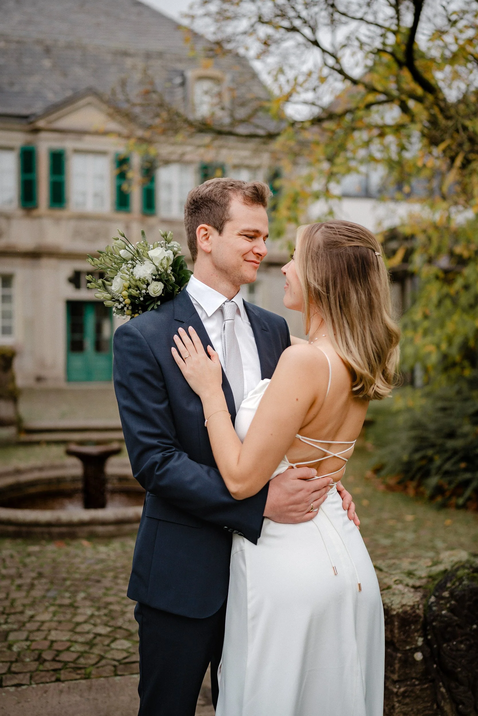 Brautpaar |Standesamtliche Hochzeit Königswinter |Fabian Dick Hochzeitsfotograf & Videograf Ein Hochzeitsfoto eines Paares, das sich liebevoll umarmt, im Garten vor einem historischen Gebäude mit Bäumen und Springbrunnen.