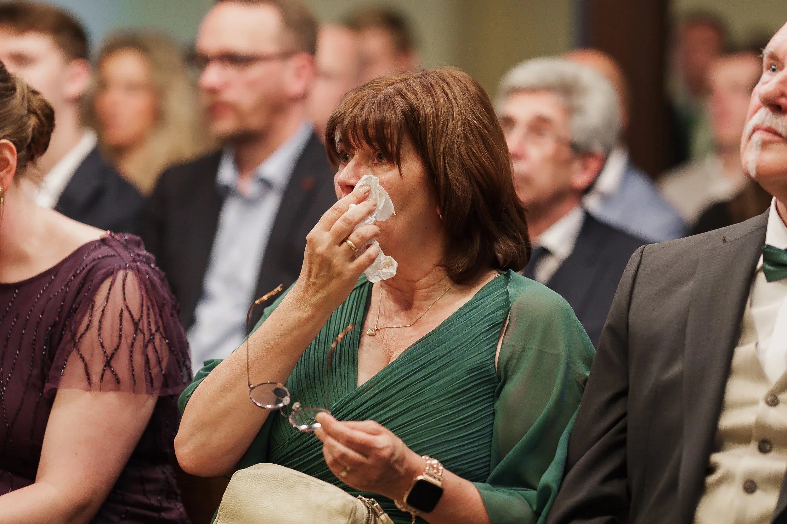 Eine Frau in einem grünen Kleid sitzt in einer Menschenmenge, wischt sich die Nase mit einem Taschentuch und hält eine Brille in der Hand.