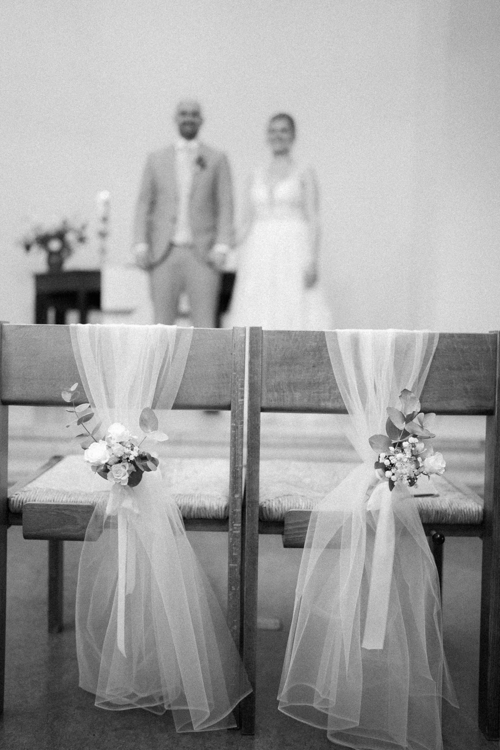 Zwei Stühle mit weißen Tüllübergängen und Blumen, im Hintergrund ein verschwommenes Brautpaar bei einer Hochzeitszeremonie.