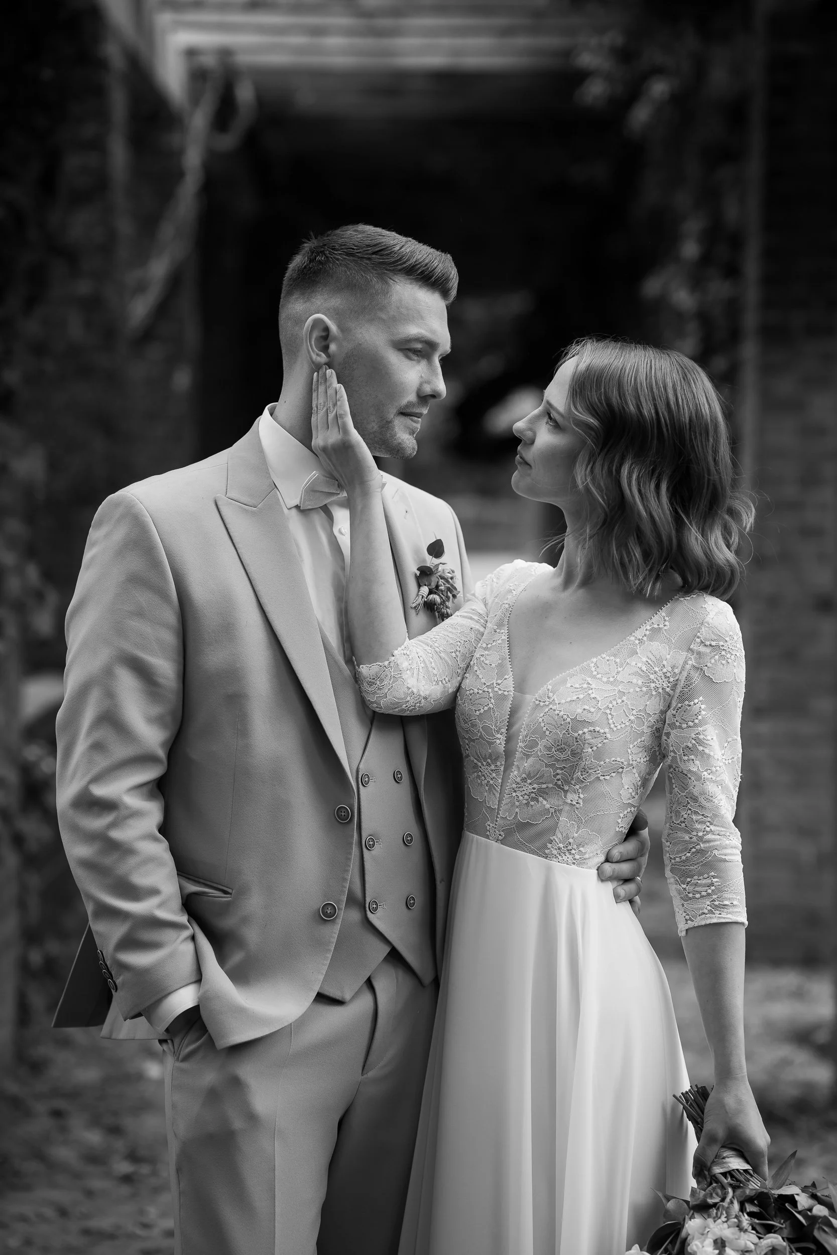Schwarz-Weiß-Foto eines Paares bei einer Hochzeit. Der Mann trägt einen Anzug mit Weste, die Frau ein langes, elegantes Brautkleid mit Spitze. Sie berühren sich zärtlich, die Frau hält einen Blumenstrauß in der Hand.