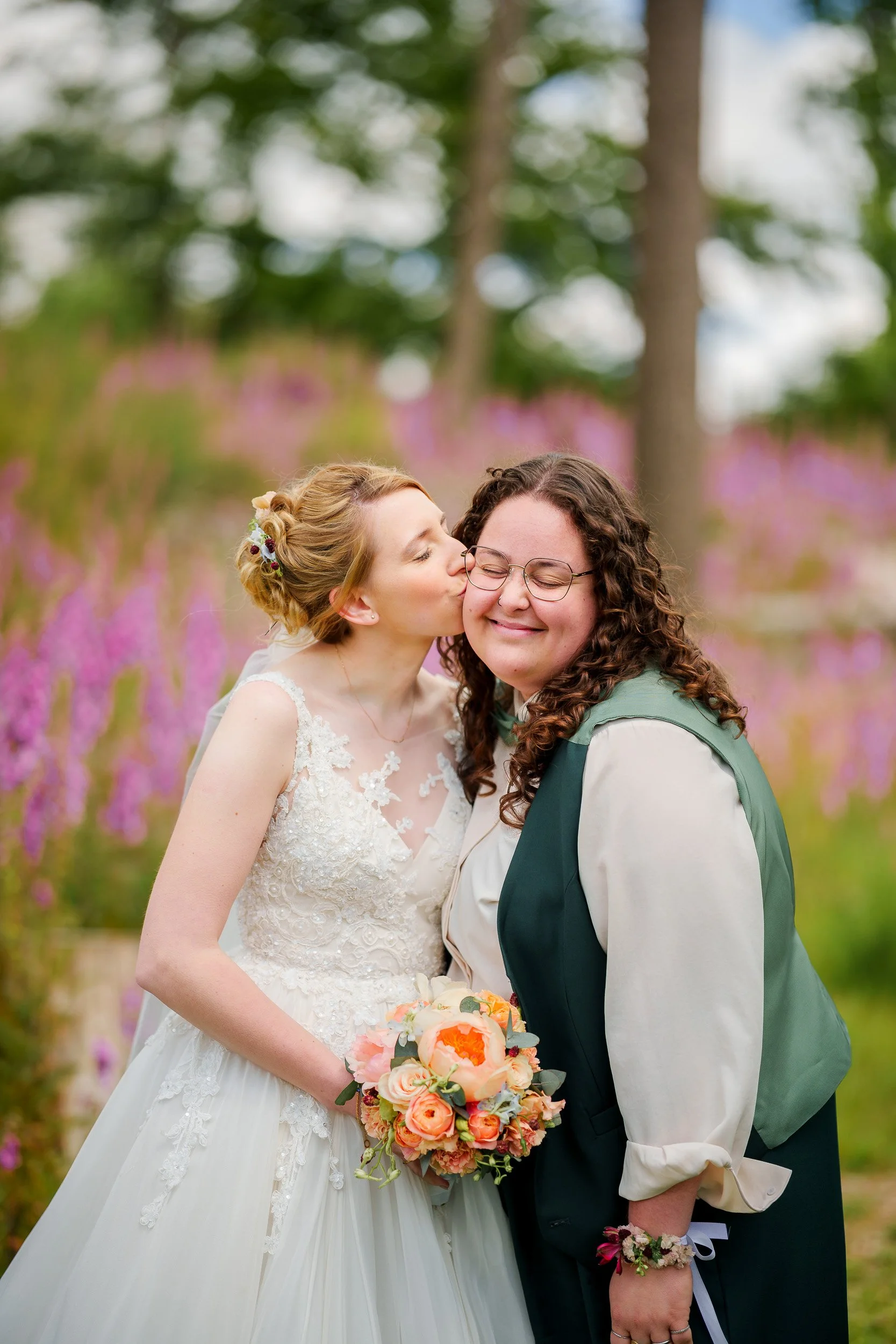 Eine Braut küsst eine andere Frau auf die Wange während einer Hochzeit im Freien, mit buntem Blumenstrauß, umgeben von pinken Blumen und Bäumen.