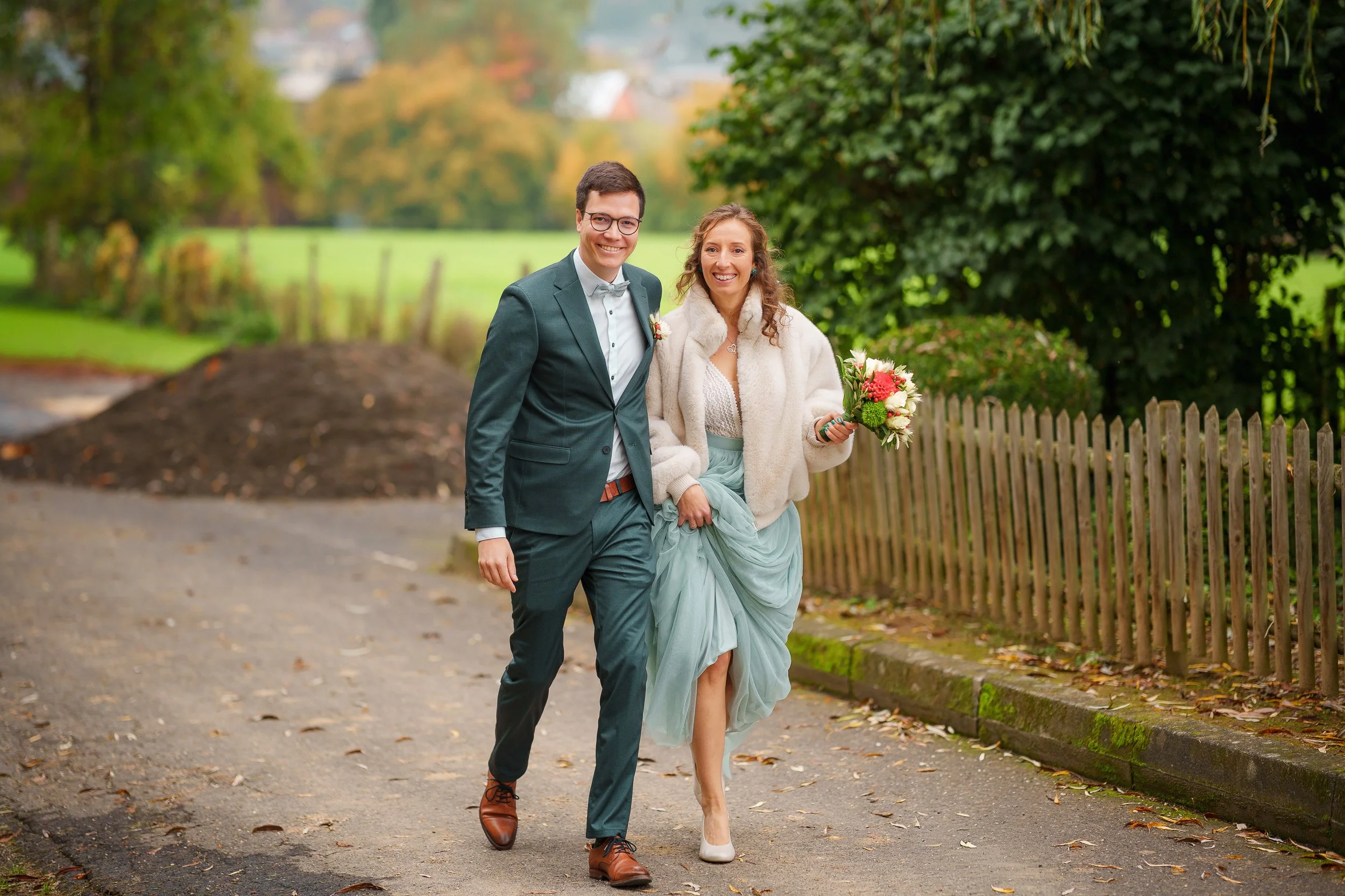 Ein Paar bei einer Hochzeit im Freien, beide lächelnd, der Mann in einem dunkelgrünen Anzug, die Frau in einem hellblauen Kleid und einer cremefarbenen Jacke, hält einen Blumenstrauß, die Umgebung ist grün mit Bäumen und einem Holzzaun.