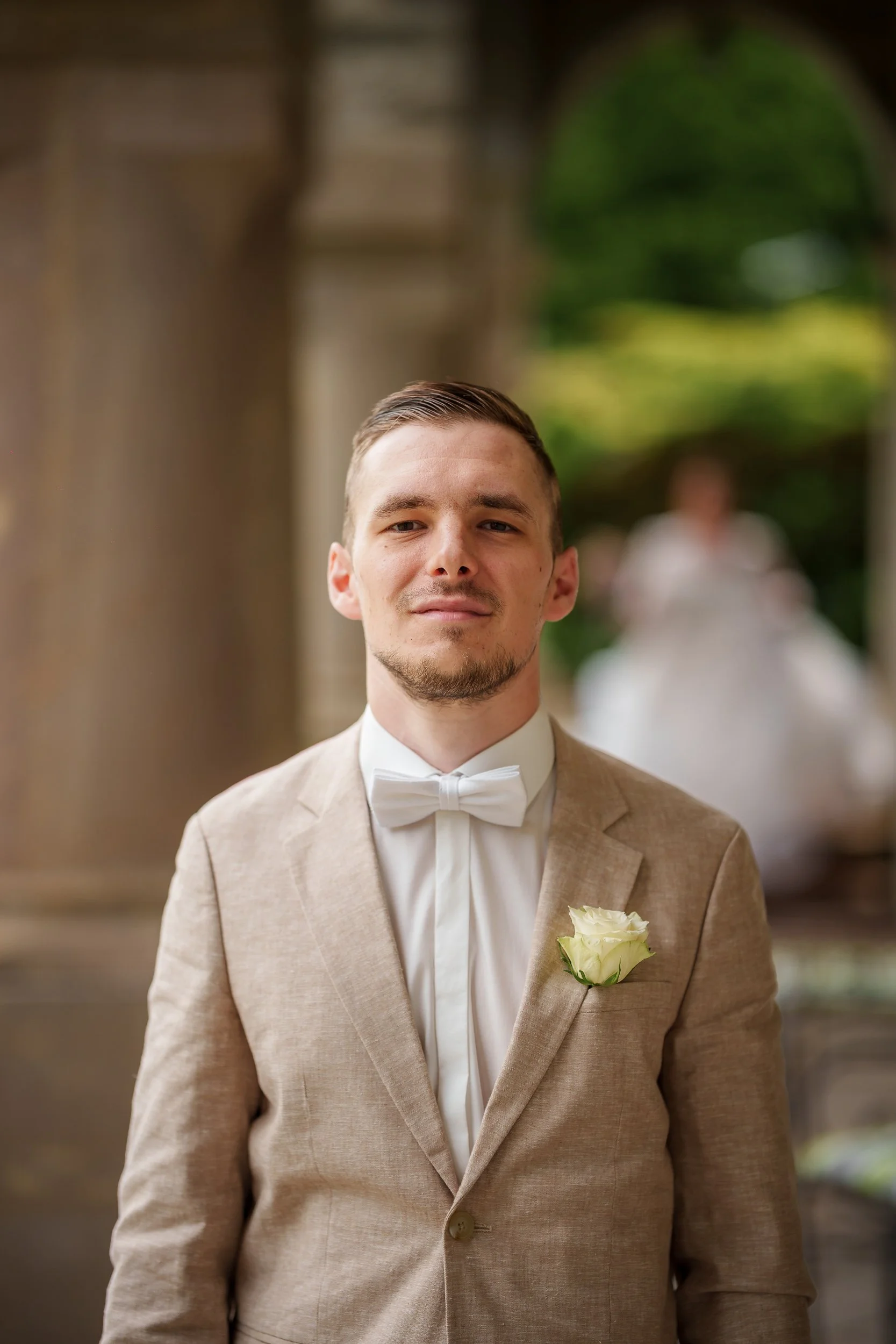 Junger Mann in beige Anzug mit weißem Hemd und Fliege, mit weiße Rose im Revers, vor einer Kirche, im Hintergrund ein unscharfes Brautpaar.