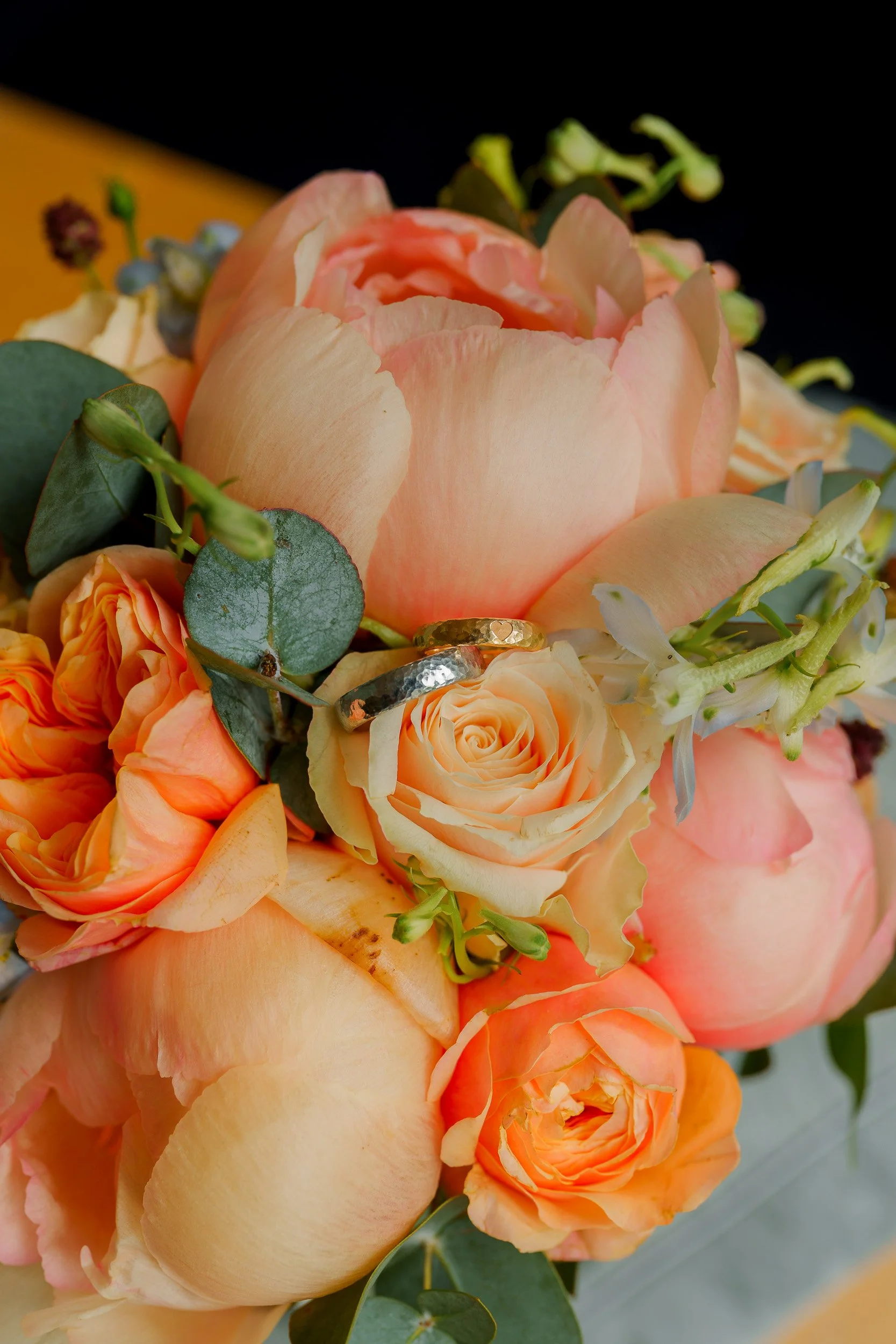 Ringshots | Nahaufnahme eines Blumenstraußes mit Pfirsichfarbenen Rosen, Eukalyptusblättern und weiteren Blumen, mit einem Ehering zwischen den Blüten.