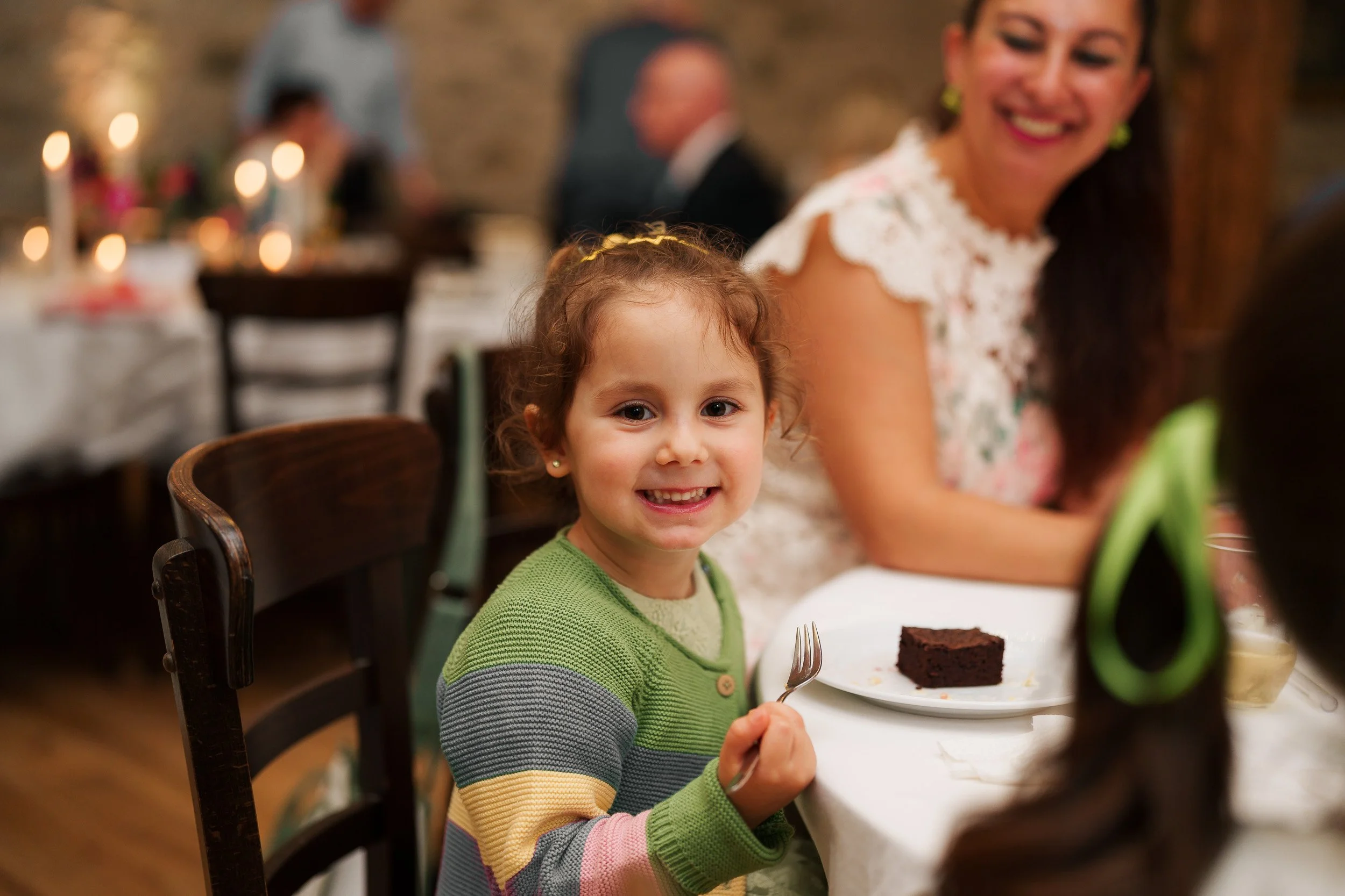 Ein lächelndes junges Mädchen mit braunen Haaren, in einem grünen, gestrickten Pullover, sitzt an einem Esstisch und hält eine Gabel. Im Hintergrund sind weitere Personen, eine Frau mit langen braunen Haaren in einer weißen, floralen Bluse und ein St