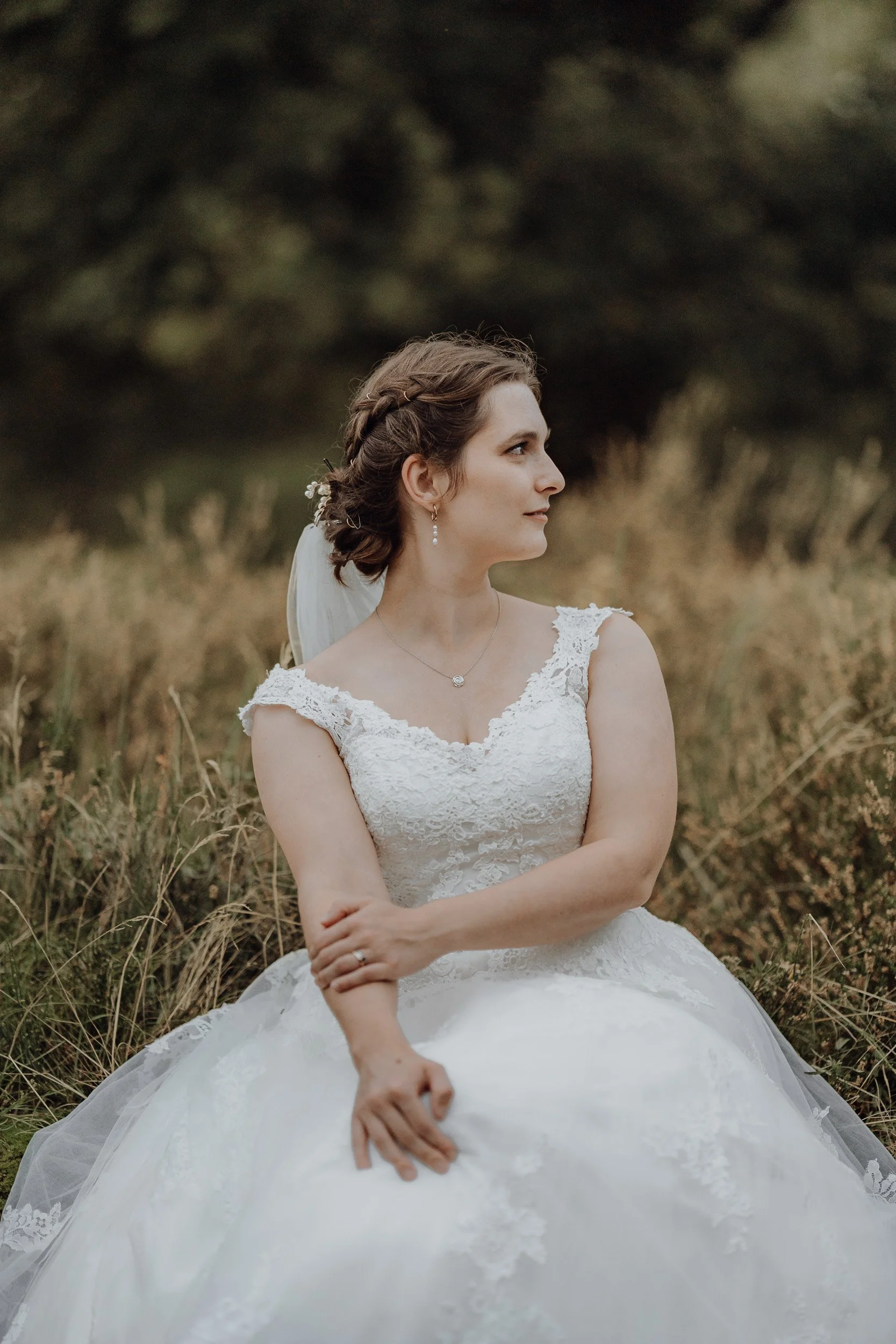 Eine Braut in einem weißen Brautkleid sitzt im Freien auf dem Gras, Blick seitwärts, mit einem grünen, bewaldeten Hintergrund.