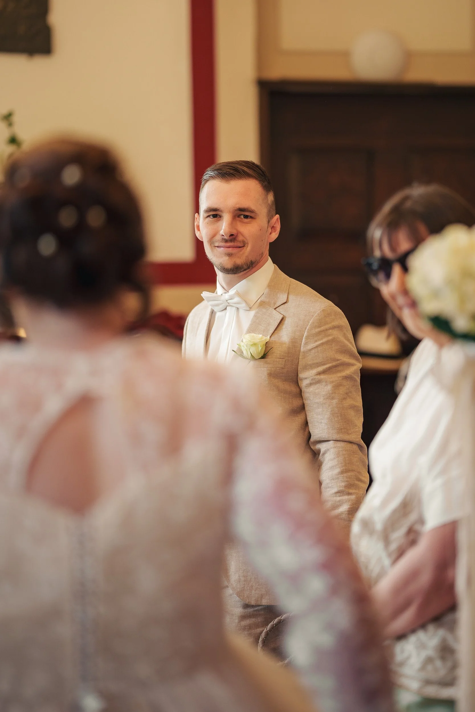 Ein Mann in einem beige Anzug und weißem Hemd mit Fliege steht bei einer Hochzeit, umgeben von Frauen, die sich lächelnd unterhalten.
