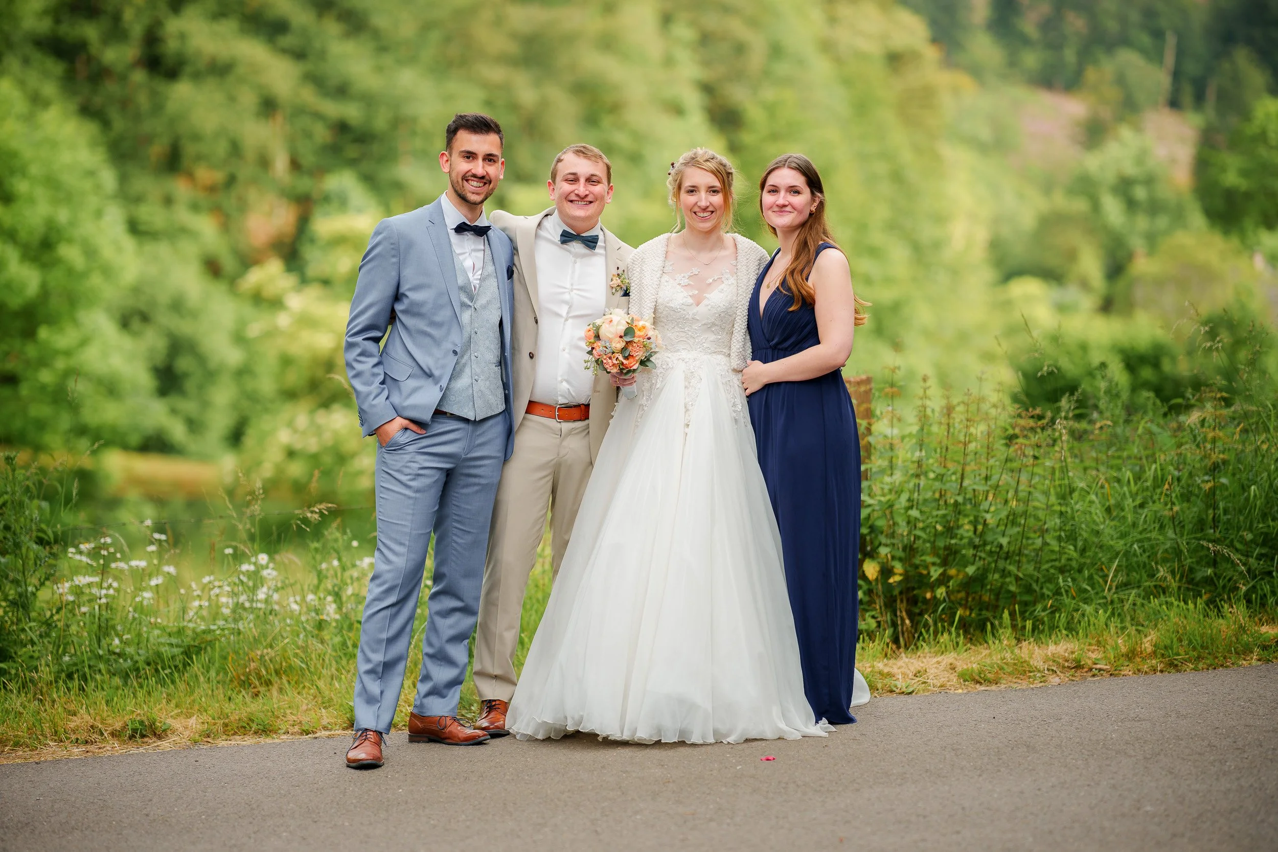 Vier Menschen auf einer Wiese, eine Hochzeitsgesellschaft, mit einem Fluss und Bäumen im Hintergrund. Ein Mann in einem blauen Anzug, ein Mann in einem beige Anzug, eine braut in einem weißen Kleid und eine Frau in einem dunklen blauen Kleid. Alle lä