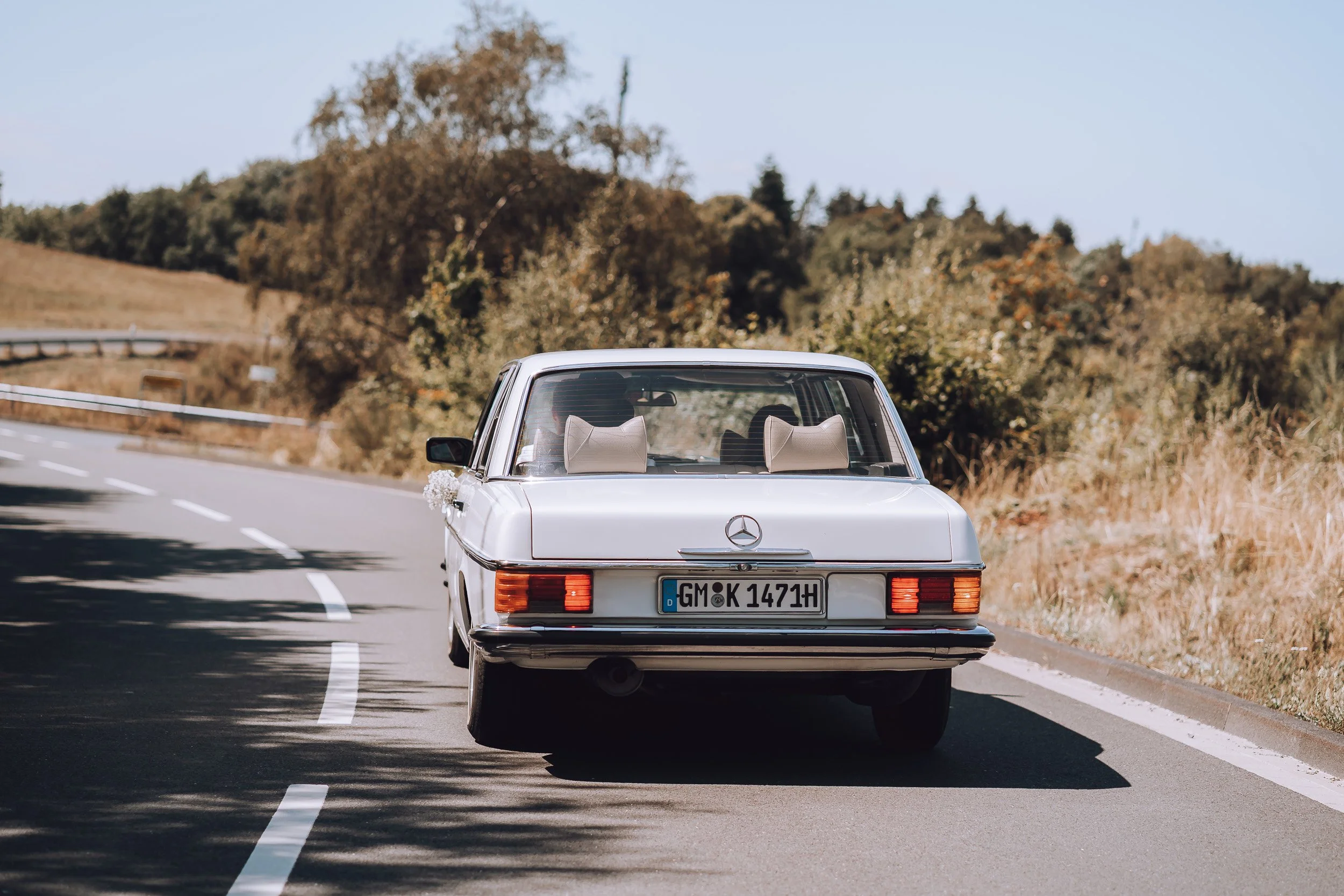 Ein weißer alter Mercedes-Benz auf einer kurvigen Landstraße mit Bäumen im Hintergrund.