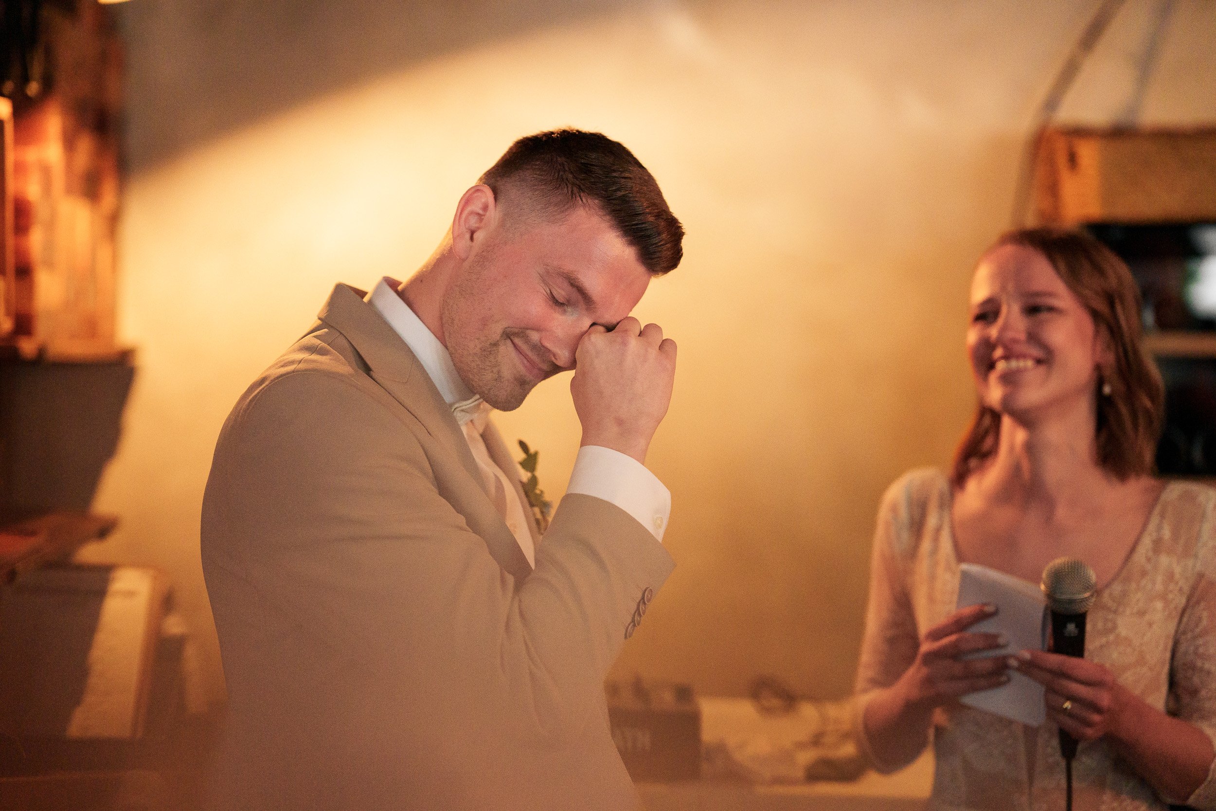 Ein Mann in einem beige Anzug ist emotional betroffen bei einer Hochzeit, während eine Frau mit einem Mikrofon ihm zu hört und lächelt.