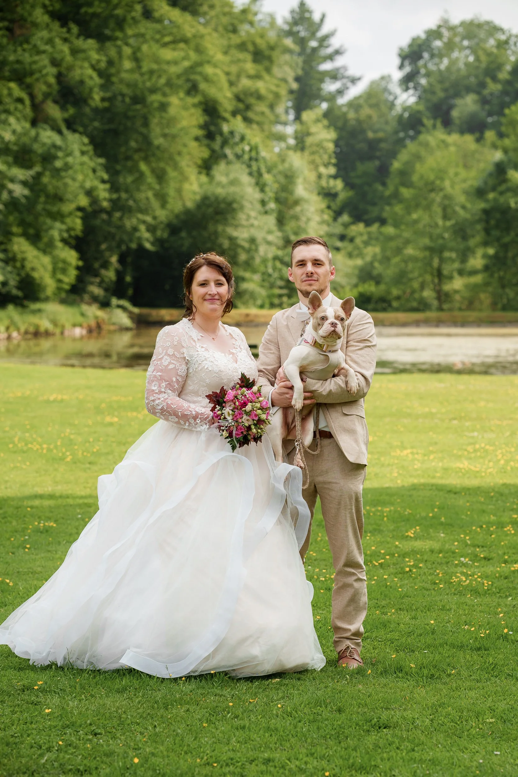 Ein Brautpaar steht im Freien auf einer grünen Wiese, begleitet von einem Hund. Die Frau trägt ein weißes Hochzeitskleid und hält einen Blumenstrauß, der Mann trägt einen beige Anzug. Im Hintergrund sind Bäume und Wasser zu sehen.