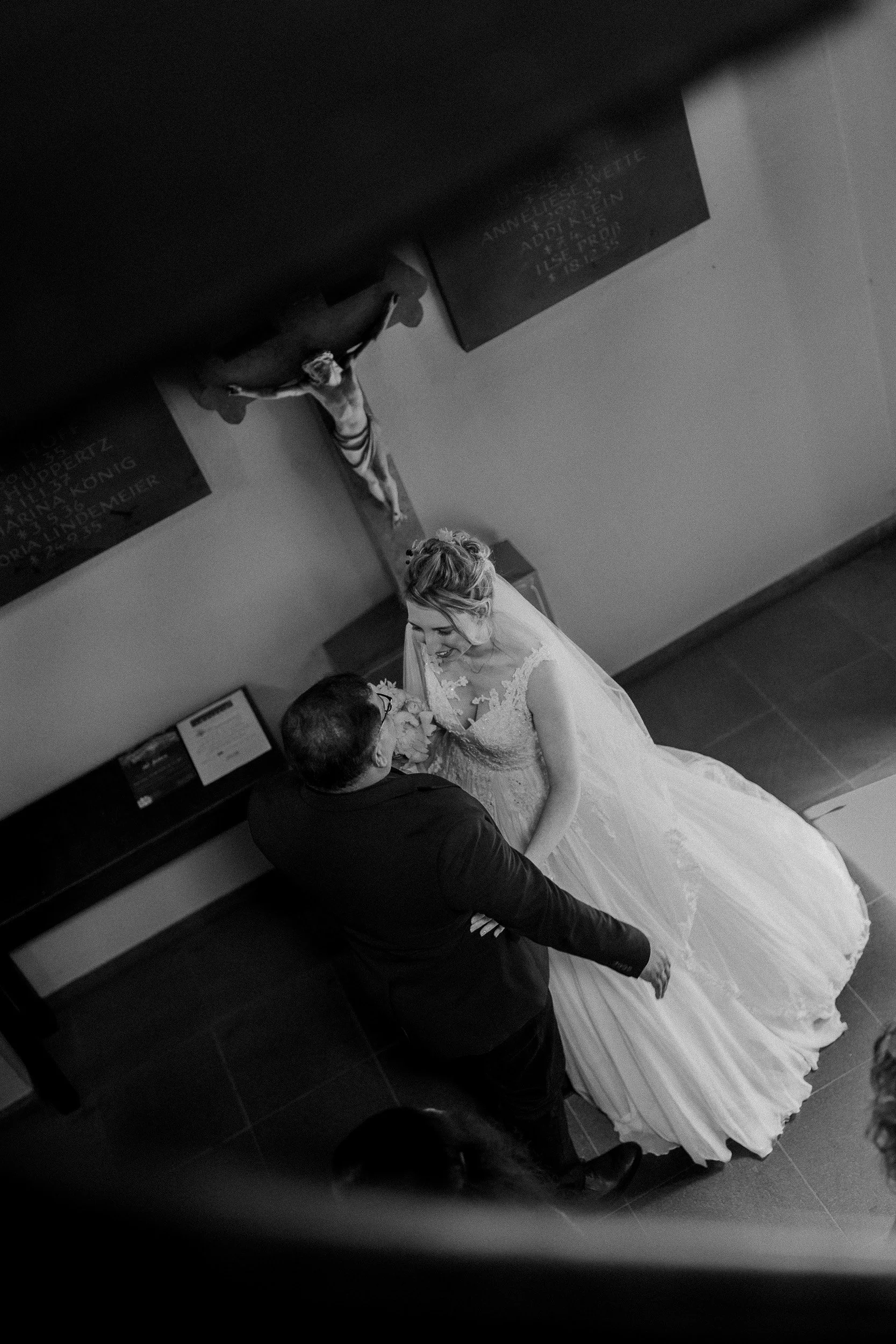 Ein Hochzeitsbild in Schwarz-Weiß, das ein Brautpaar zeigt, das sich liebevoll ansieht, während sie tanzen. Der Hintergrund enthält eine Wand mit einem Kreuz und einer Tafel, sowie einen Mann, der etwa durch den Raum geht.