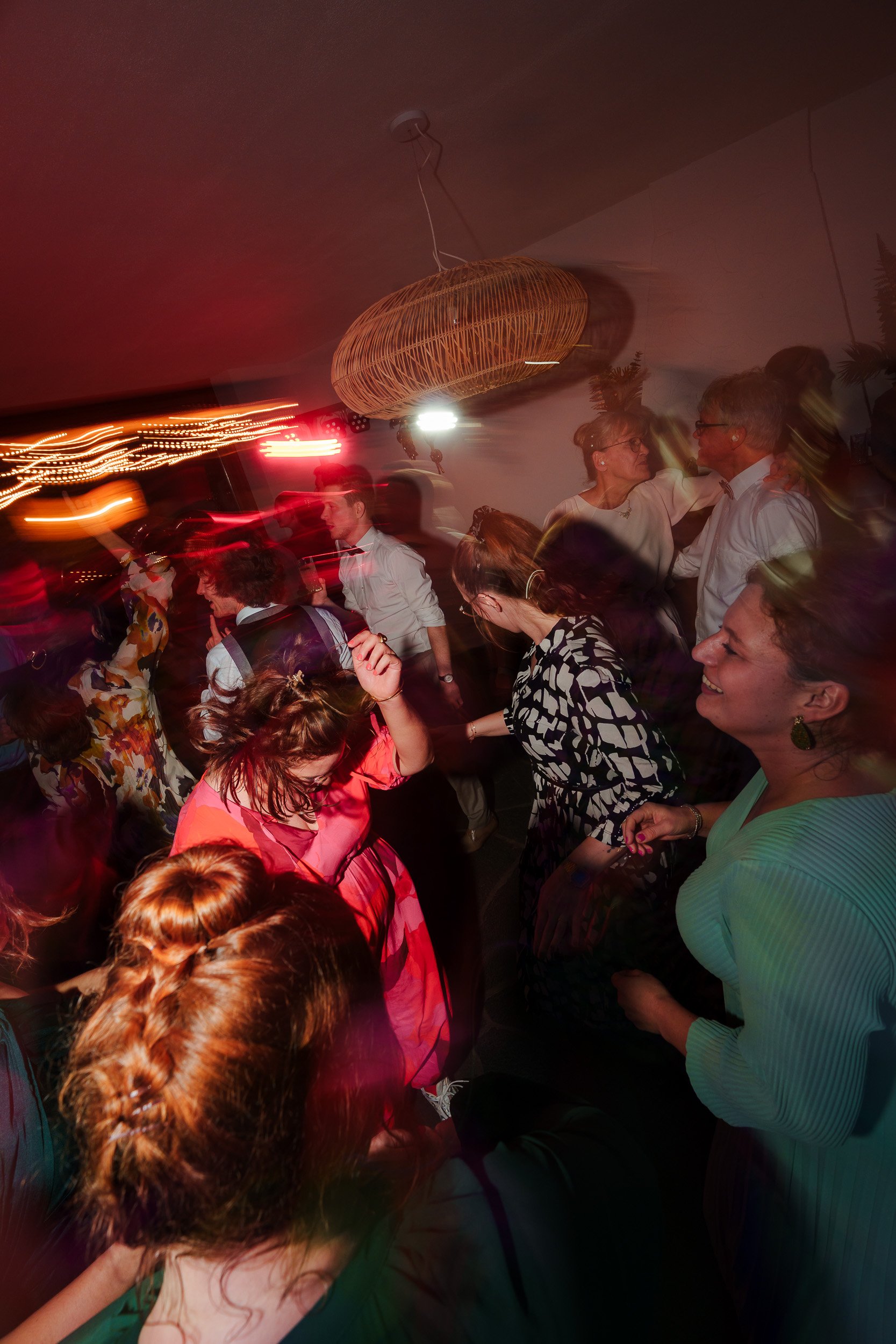Menschen tanzen und feiern bei einer Party in einem dimmigen Raum mit bunten Lichtern und einem runden, geflochtenen Lampenschirm an der Decke.