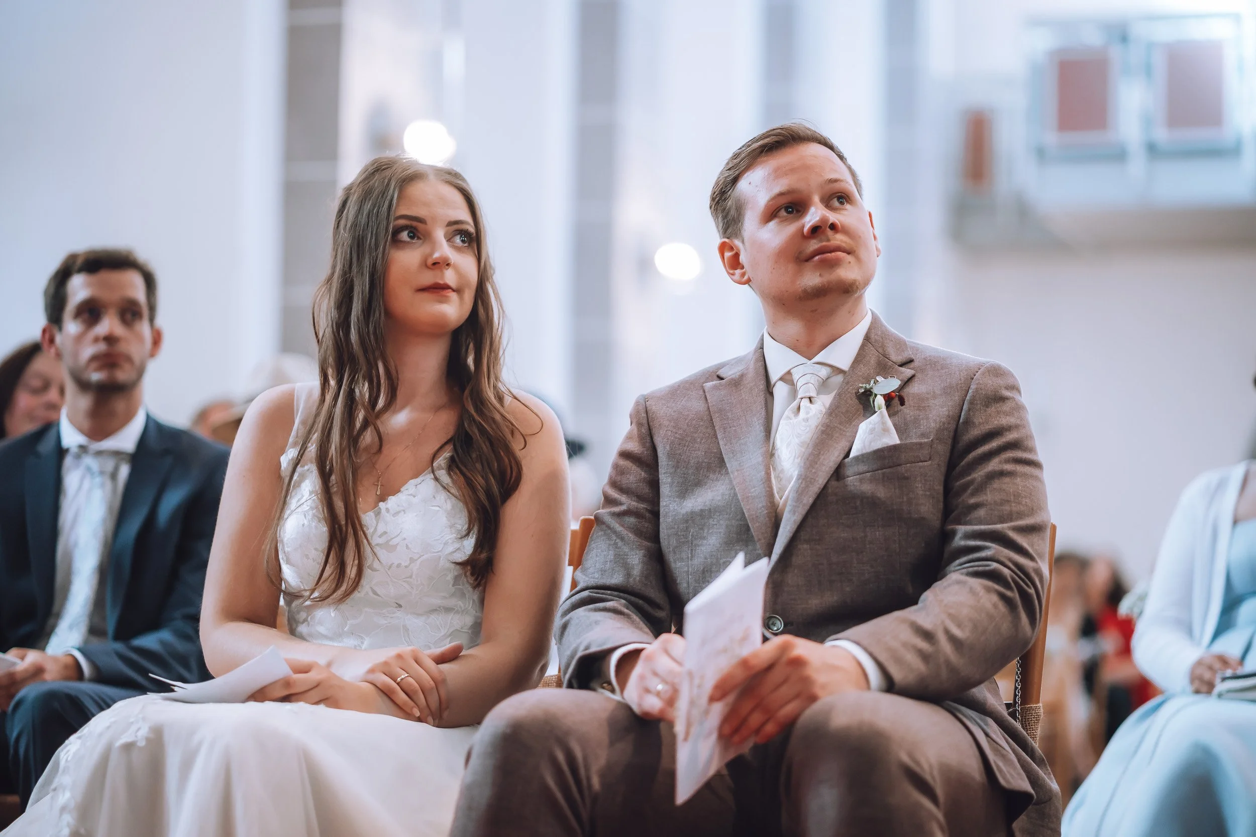 Hochzeitspaar sitzt in einer Kirche bei der Zeremonie, alle blicken nach vorne, die Frau trägt ein weißes Kleid, der Mann einen grauen Anzug.