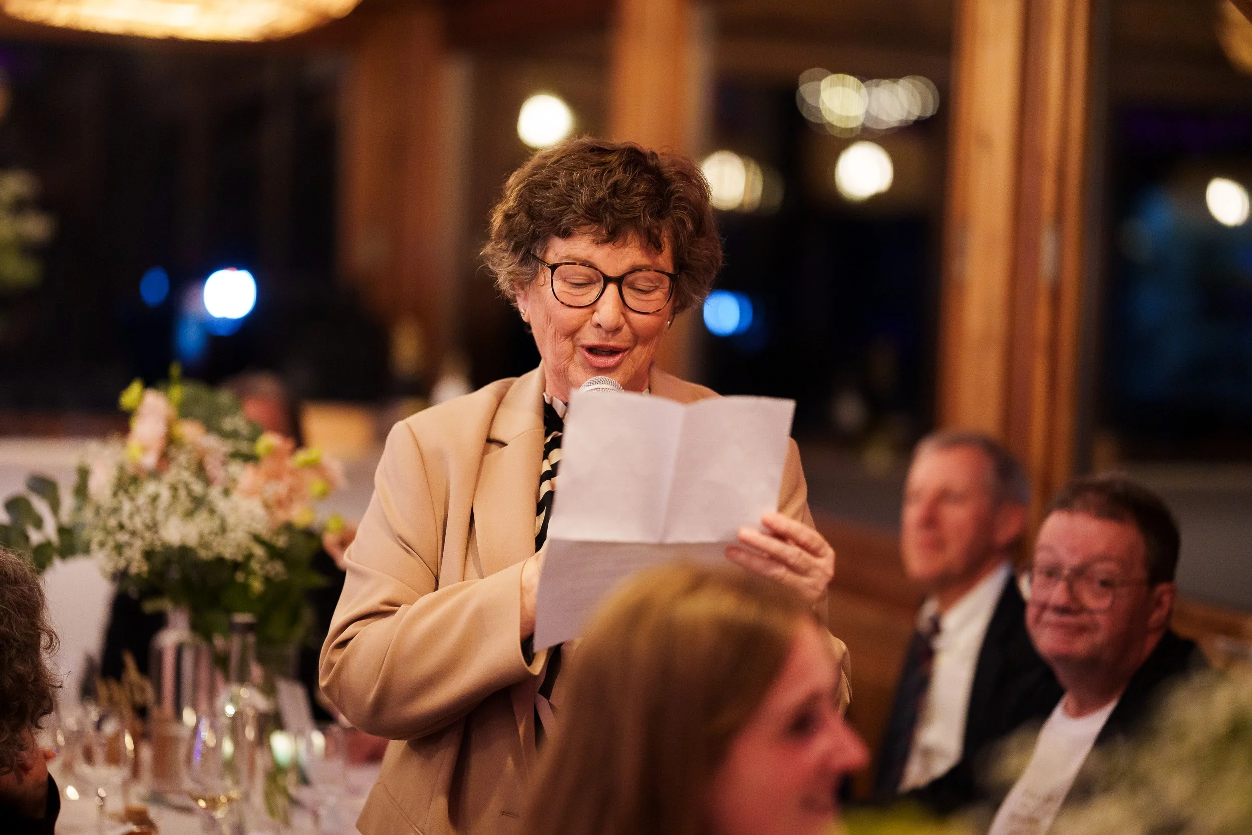 Eine ältere Frau liest auf einer Hochzeitsfeier eine Rede vor einer Gruppe von Gästen, die an einem festlich gedeckten Tisch sitzen.