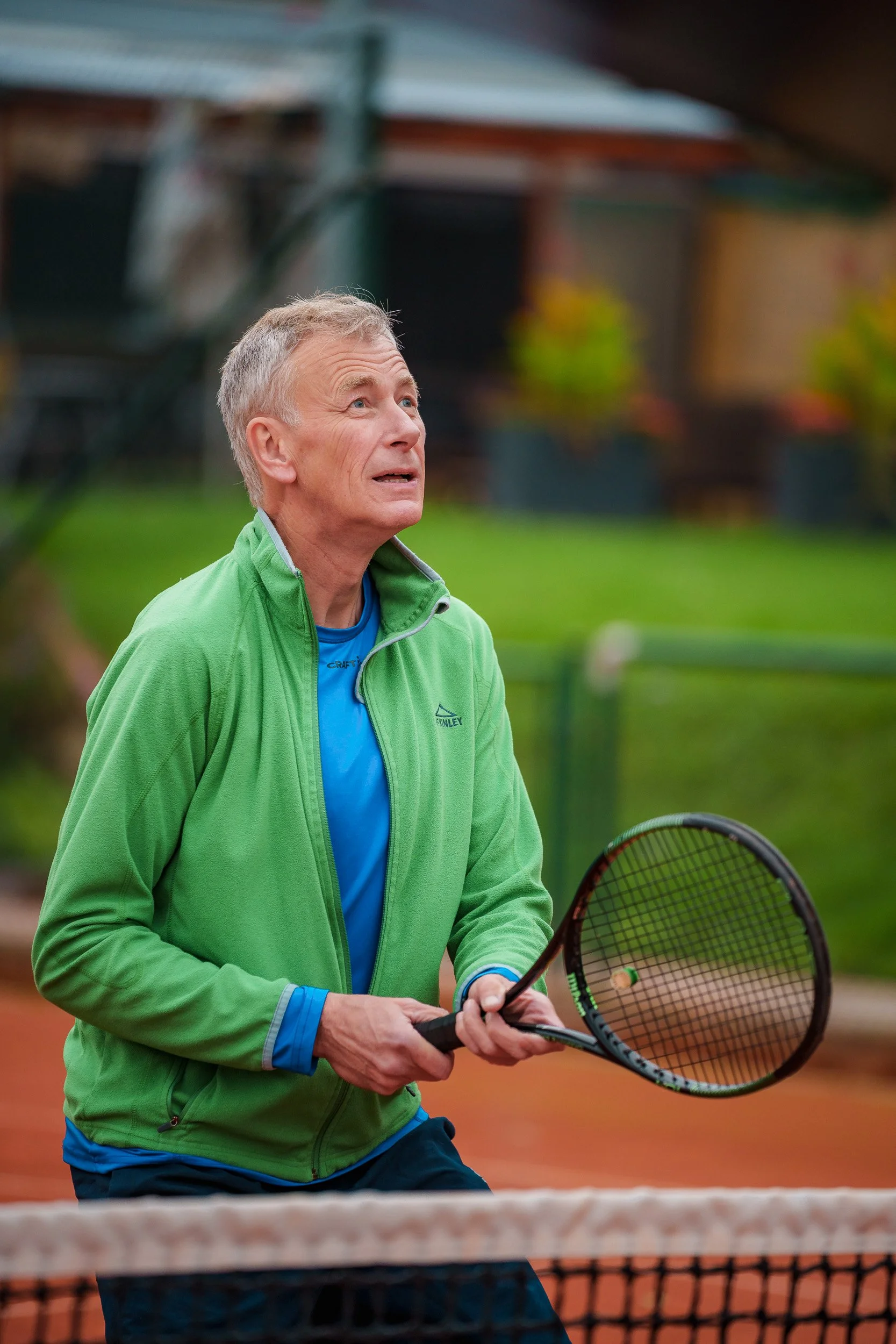 Ein älterer Mann spielt Tennis auf einem Freiplatz, trägt eine grüne Jacke und hält einen Tennisball sowie einen Schläger.