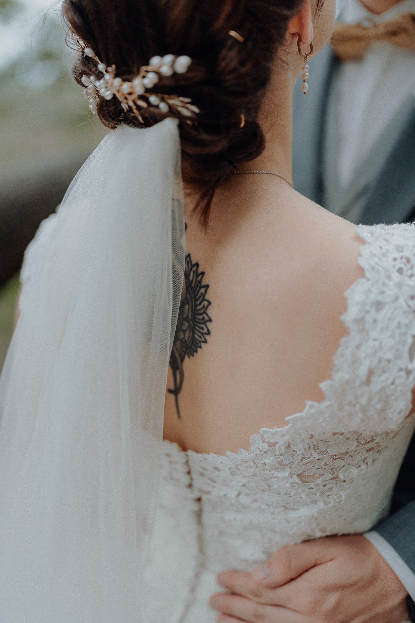 Nahaufnahme eines Brautpaares bei ihrer Hochzeit, Fokus auf die Braut mit weißen Spitzenkleid, Schleier und einer Tätowierung auf der Schulter, die Hand des Bräutigams liegt auf ihrer Taille.