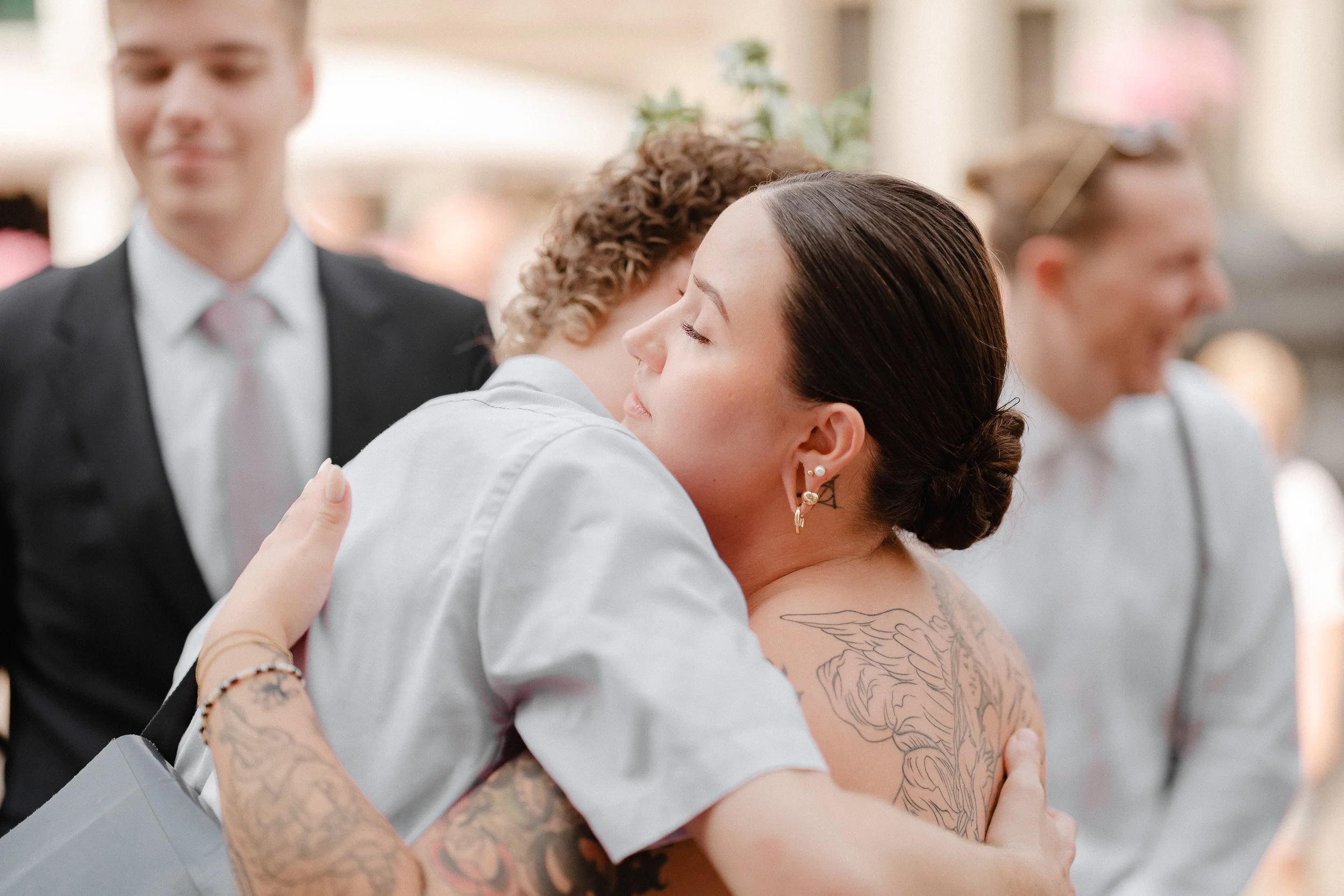 Zwei Frauen umarmen sich herzlich bei einer Feier, im Hintergrund sind weitere Personen im formellen Dress, die sich freuen.