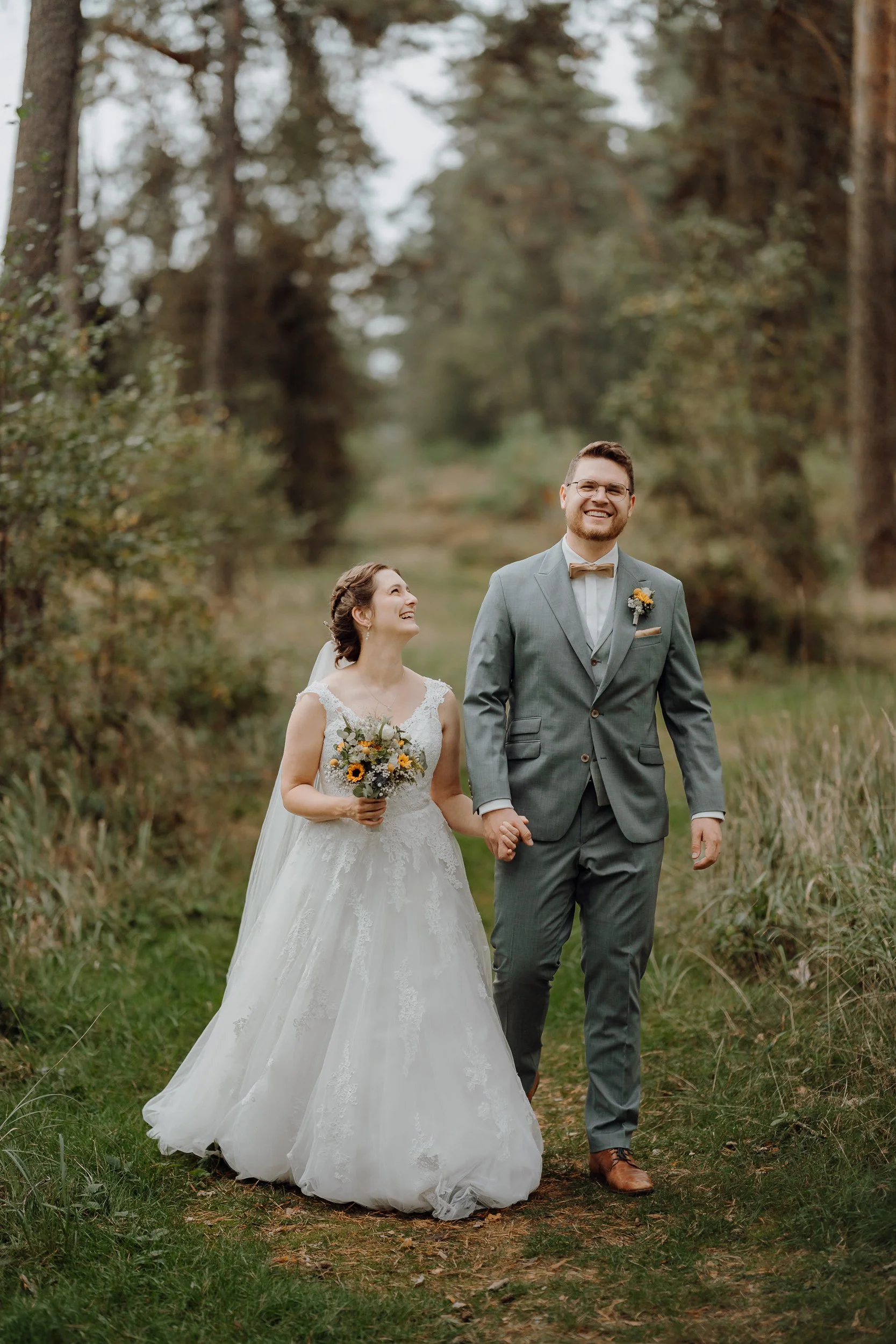 Ein Brautpaar bei einem Spaziergang im Wald, lächelnd und Hand in Hand, im Hochzeitsoutfit, umgeben von Bäumen und Natur.