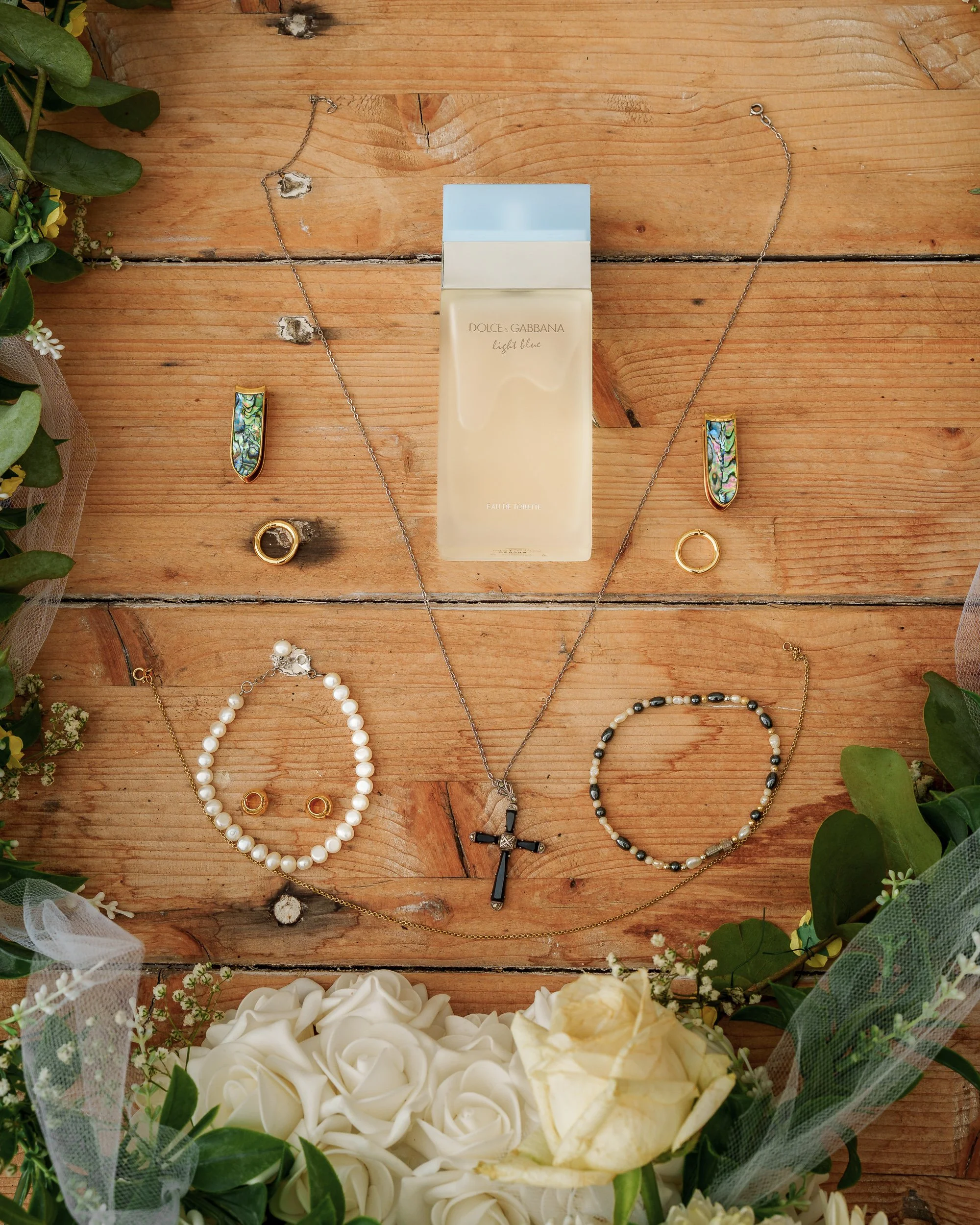Verschiedene Schmuckstücke, darunter Ringe, Halsketten, Ohrringe und Armbänder, sind auf einem Holztisch mit einem Parfümflakon in der Mitte arrangiert, umgeben von grünen Blättern und weißen Rosen.