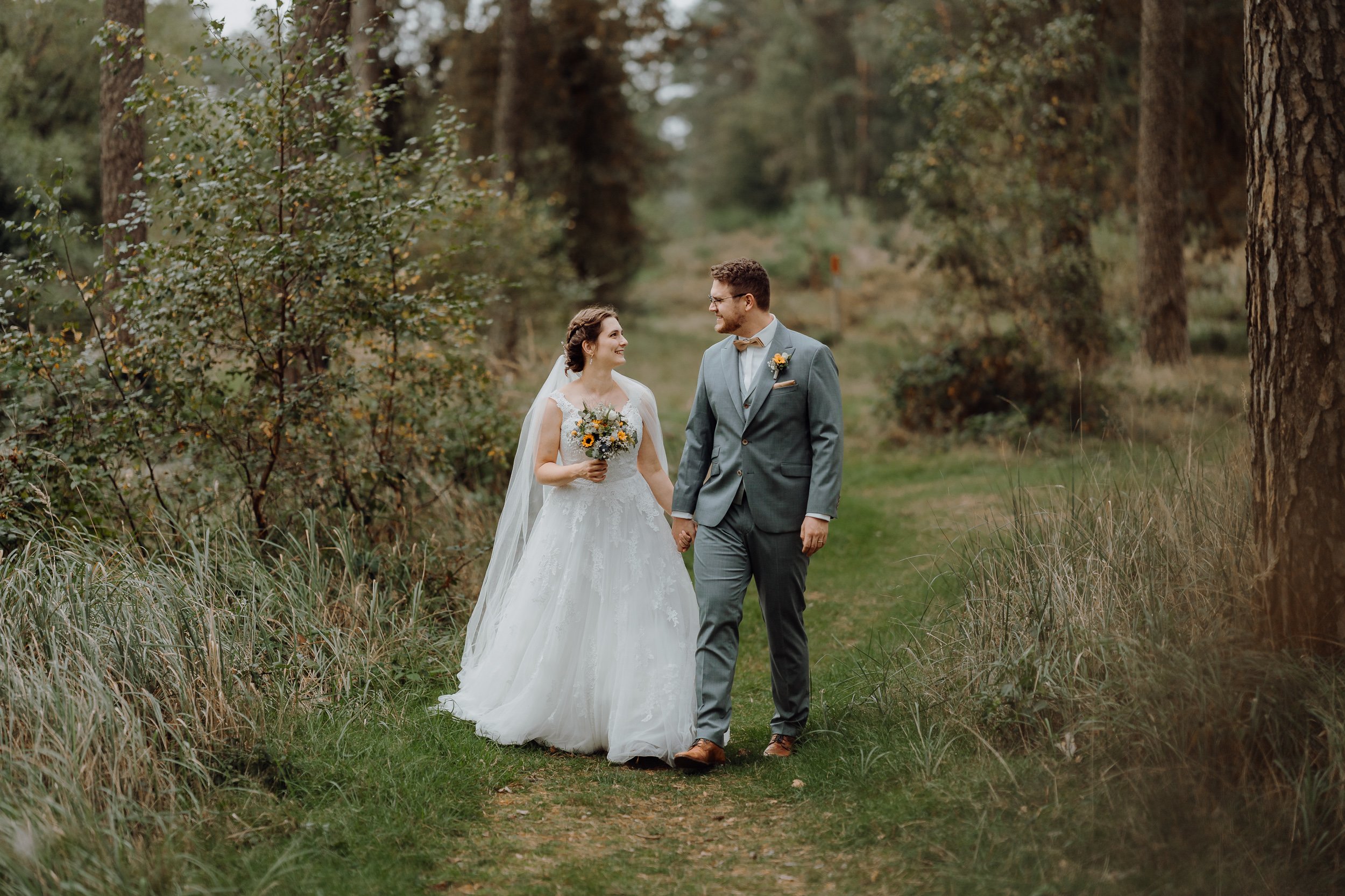Ein Brautpaar in der Natur, die Braut trägt ein weißes Hochzeitskleid mit Schleier, der Bräutigam trägt einen grauen Anzug mit Fliege, sie halten sich an den Händen und schauen sich an.