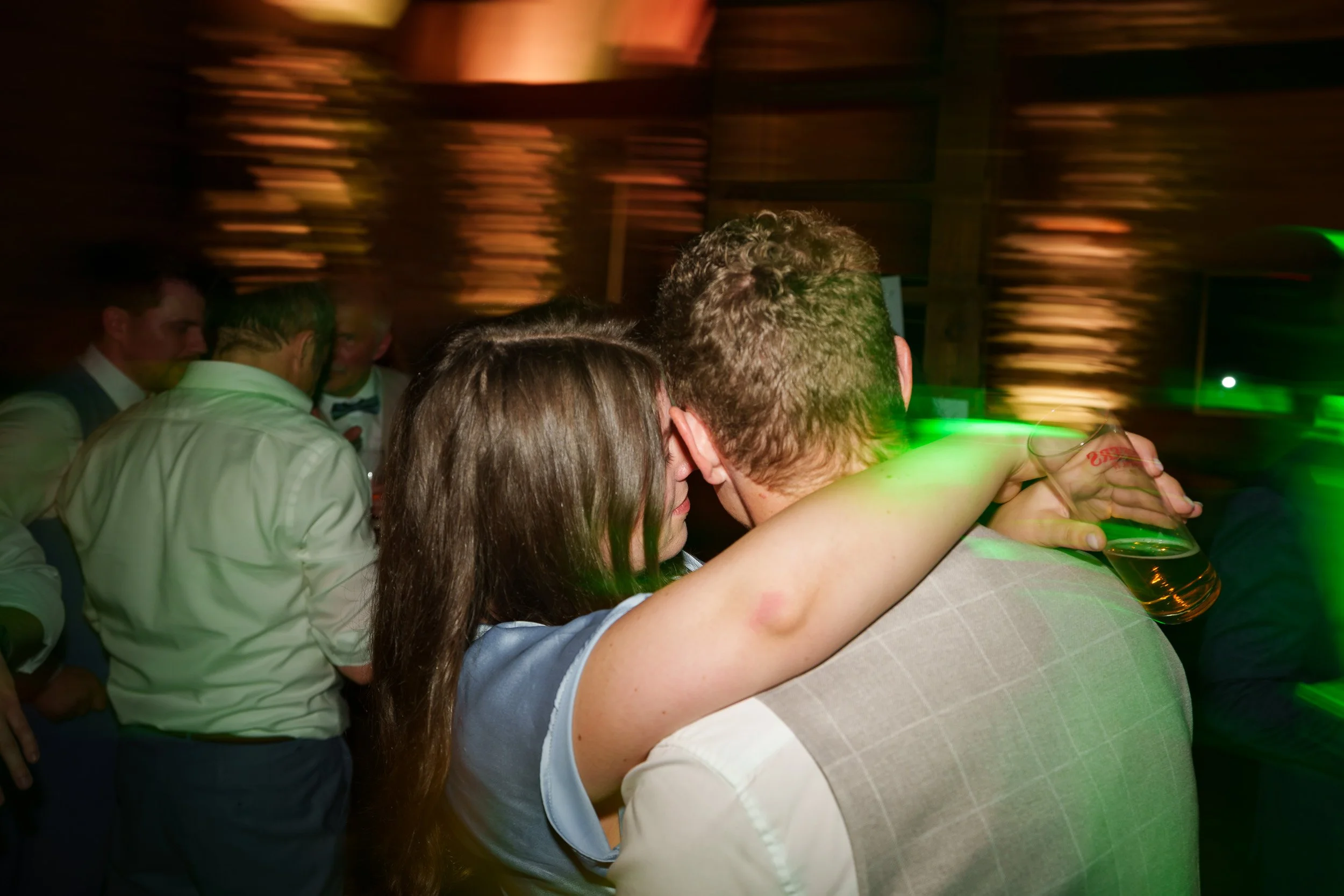 Ein Paar tanzt eng umarmend auf einer Party, im Hintergrund weitere Gäste, bunte Licht- und Bewegungseffekte.