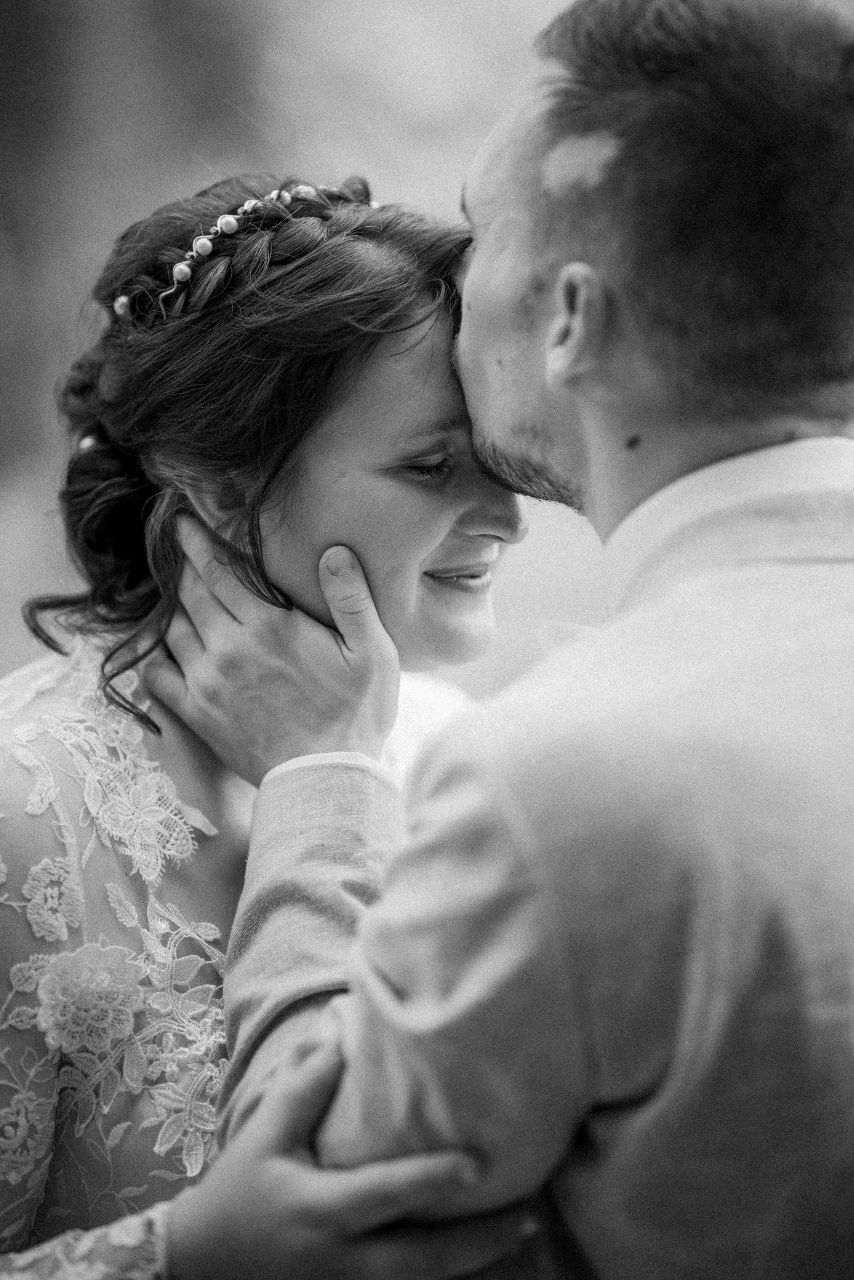 Ein hochzeitsnahes Schwarz-Weiß-Foto eines Paares, das sich liebevoll berührt, mit Blicken und Nasen, die sich fast berühren. Die Frau trägt eine Brautkleid mit floralem Spitzenmuster und hat eine Haarnadel mit Perlen im Haar. Der Mann hat dunkles, k