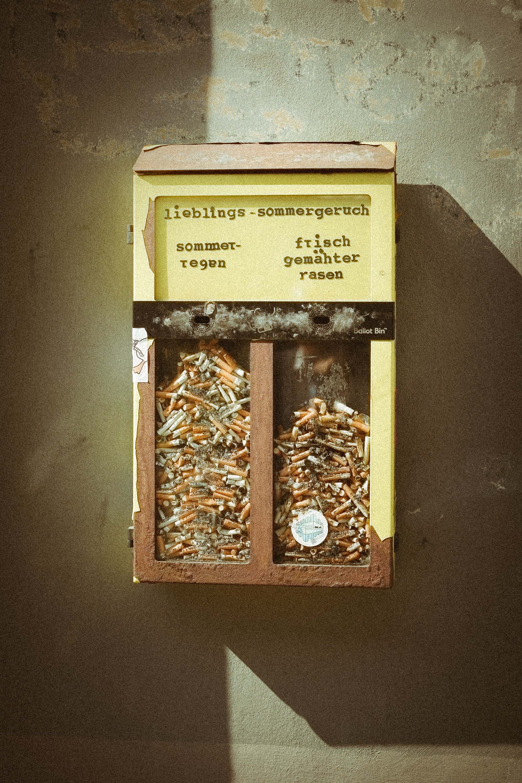 Eine alte Briefkastenbox, die mit Zigarettenstummeln und Staub gefüllt ist, mit gelbem Schild und schwarzen Aufschriften.