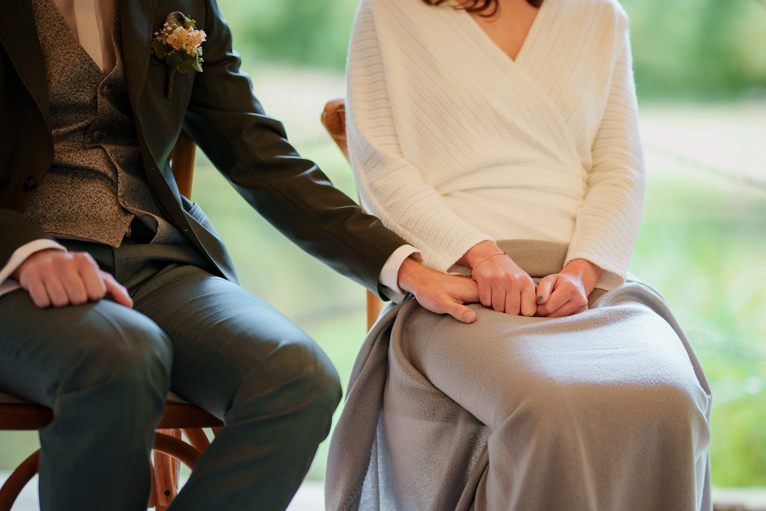 Zwei Personen halten sich an den Händen, eine Person trägt einen weißen Pullover und einen grauen Rock, die andere Person trägt einen braunen Anzug mit Blumenanstecker.