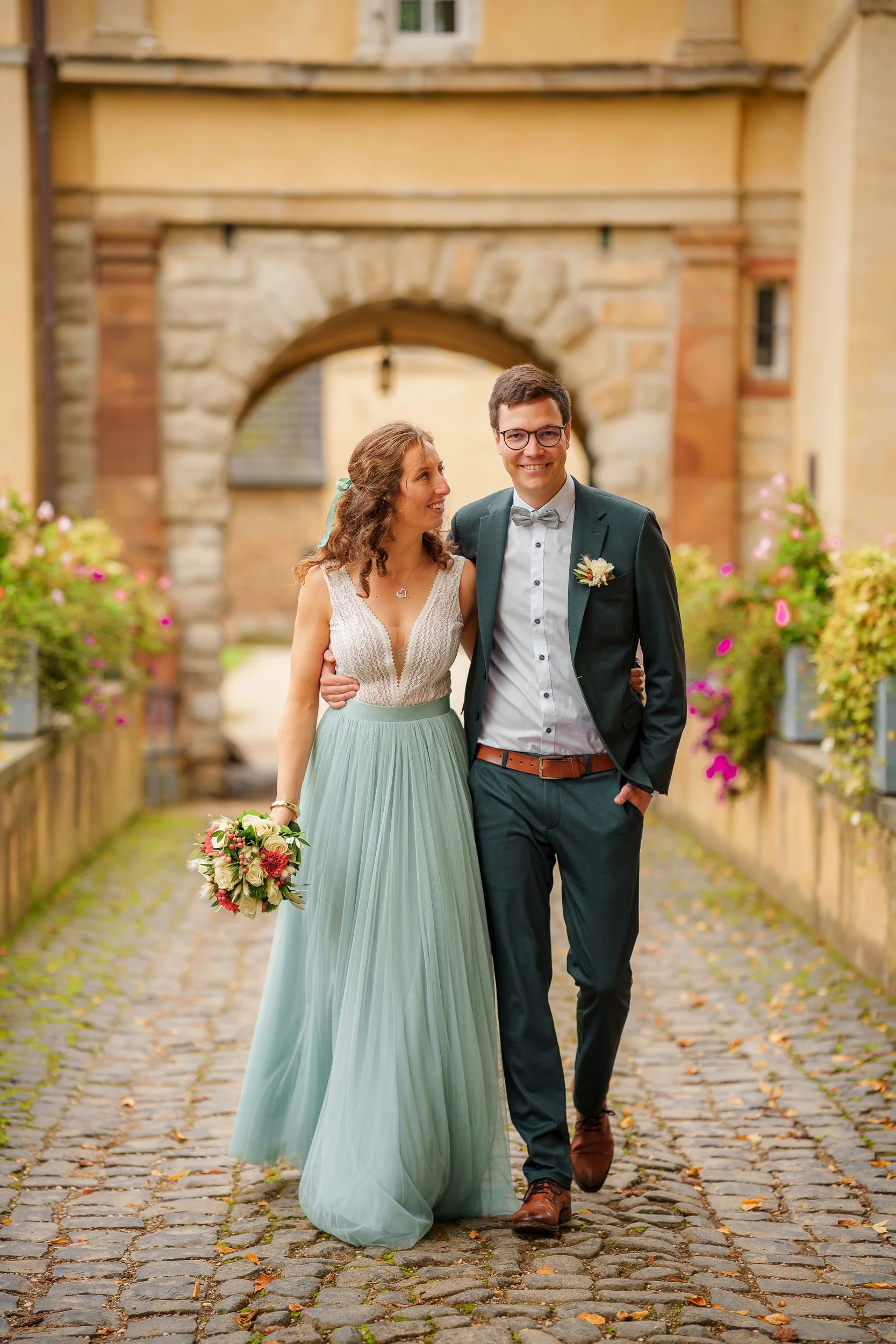 Ein Paar bei ihrer Hochzeit, das Hand in Hand durch eine Altstadtgasse geht, die mit Blumen bepflanzten Beeten flankiert ist. Die Frau trägt ein langes, grünes Brautkleid und hält einen Blumenstrauß. Der Mann trägt einen dunklen Anzug mit Fliege. Im 