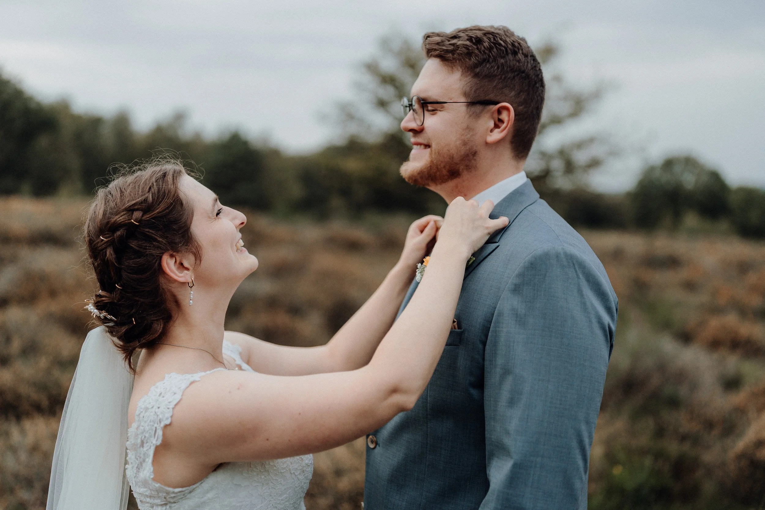 Ein Brautpaar, das sich an einem Tag im Freien umarmt, lächelnd und glücklich, die Braut trägt ein weißes Spitzenkleid und die Haare sind zu einer Hochsteckfrisur gestylt, der Bräutigam trägt einen grauen Anzug und hat eine Brille.