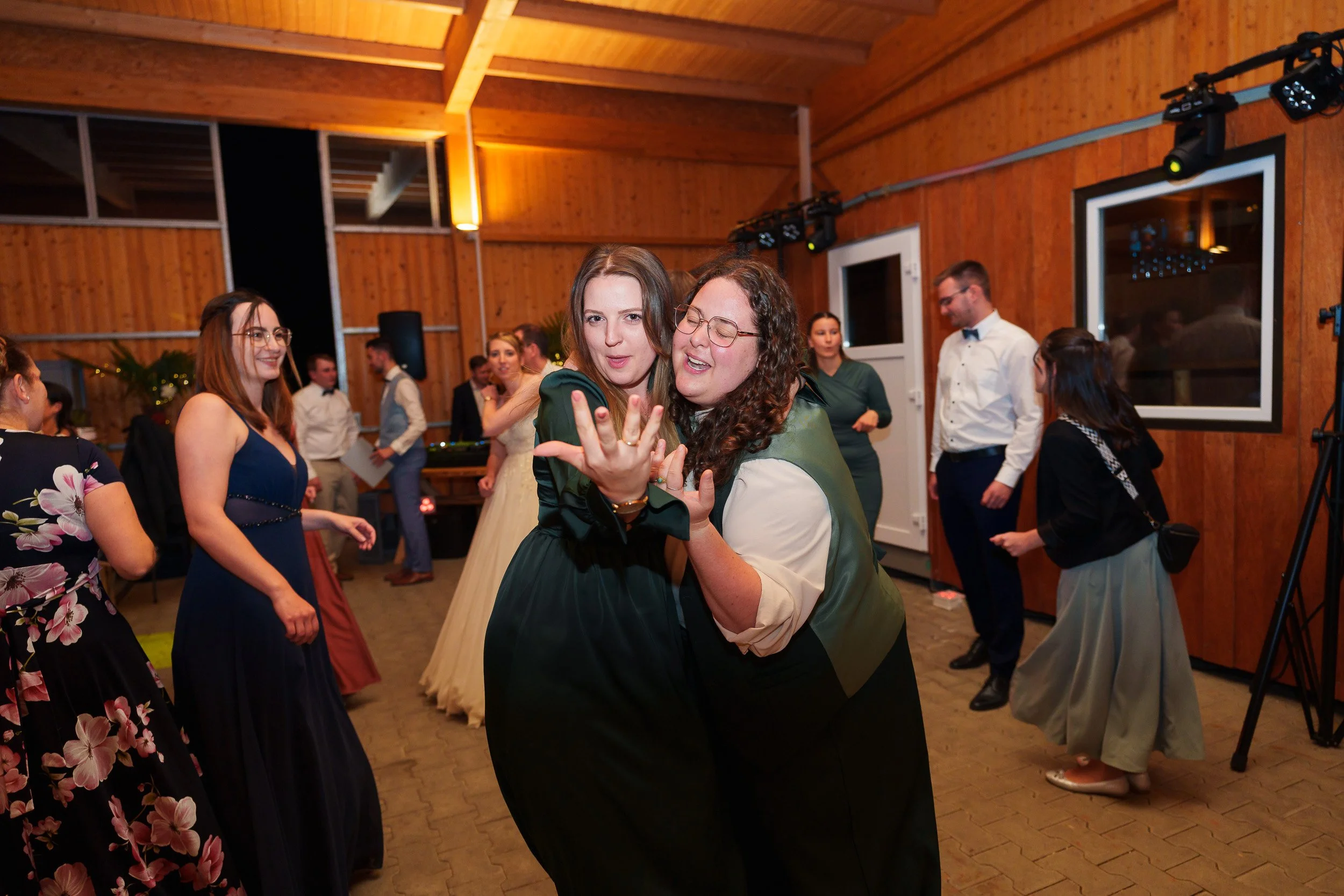 Menschen tanzen bei einer Feier in einem Holzzimmer, zwei Frauen im Vordergrund lächeln und posieren für die Kamera, während andere Gäste im Hintergrund ebenfalls tanzen und sich unterhalten.