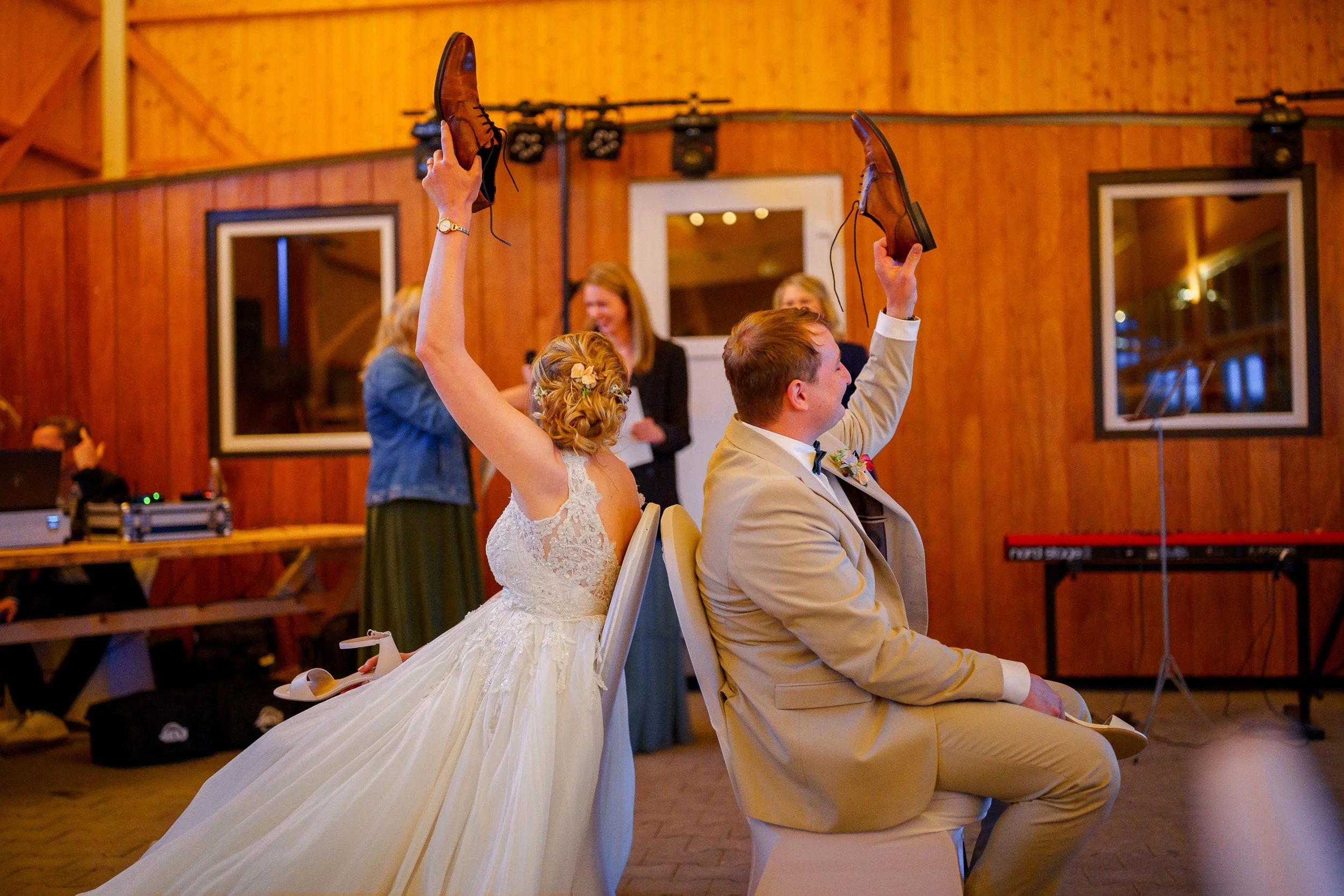 Ein Brautpaar spielt das Stuhltanz-Spiel bei einer Hochzeit. Das Brautpaar sitzt auf Stühlen, sie lehnt sich gegen ihn, beide halten ihre Schuhe in der Hand hoch.