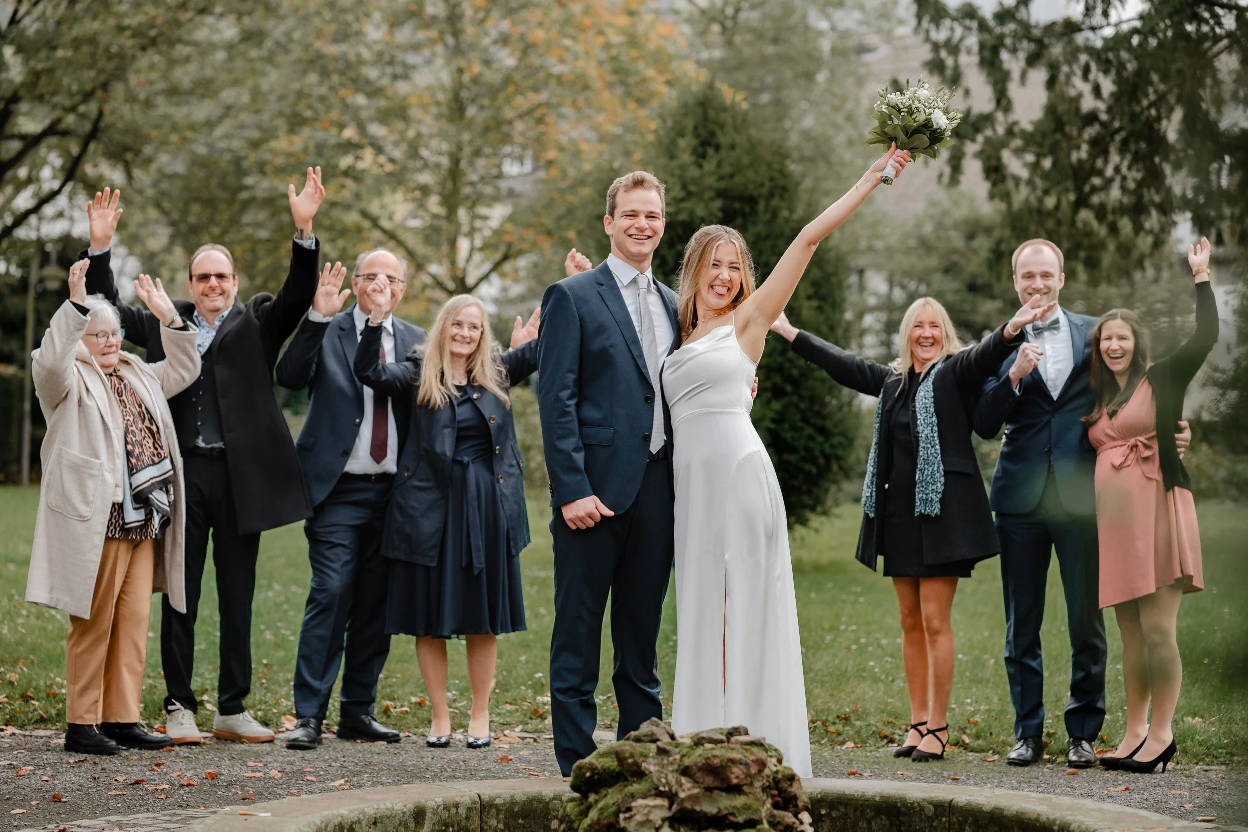 Ein Hochzeitsfoto im Freien mit lachendem Brautpaar und Gästen im Hintergrund.