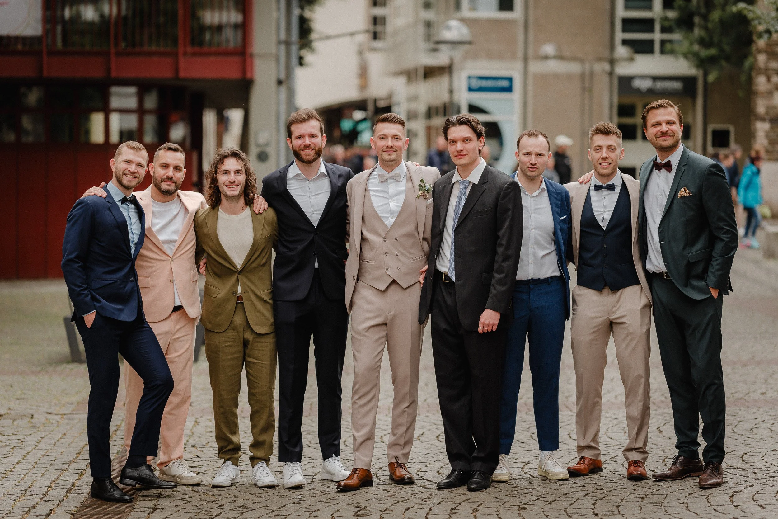 Eine Gruppe von Männern in formeller Kleidung, vermutlich beim Feiern eines besonderen Anlasses, steht auf einem gepflasterten Platz in einer Stadt.