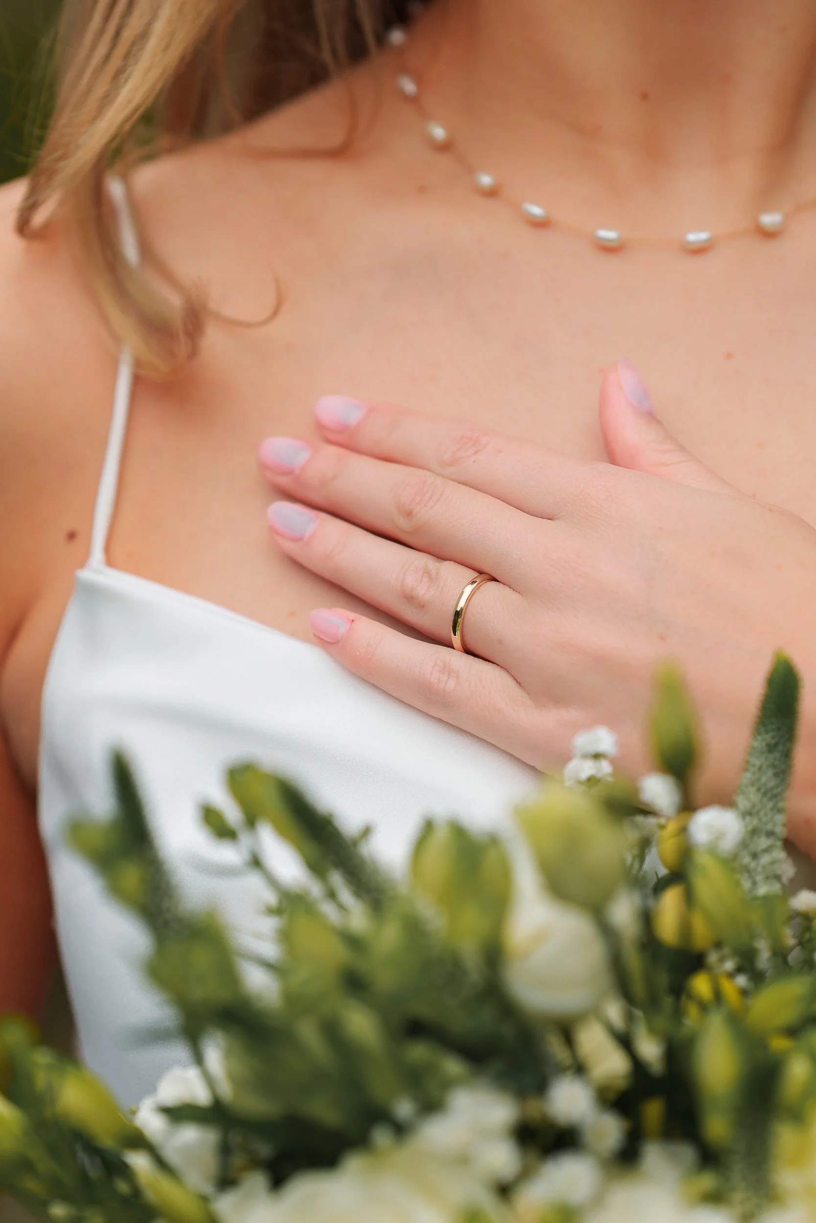Nahaufnahme einer Frau in einem weißen Kleid, die eine Hand mit einem goldenen Ehering auf die Brust legt, trägt eine Perlenkette, und im Vordergrund sind weiße Blumen sichtbar.