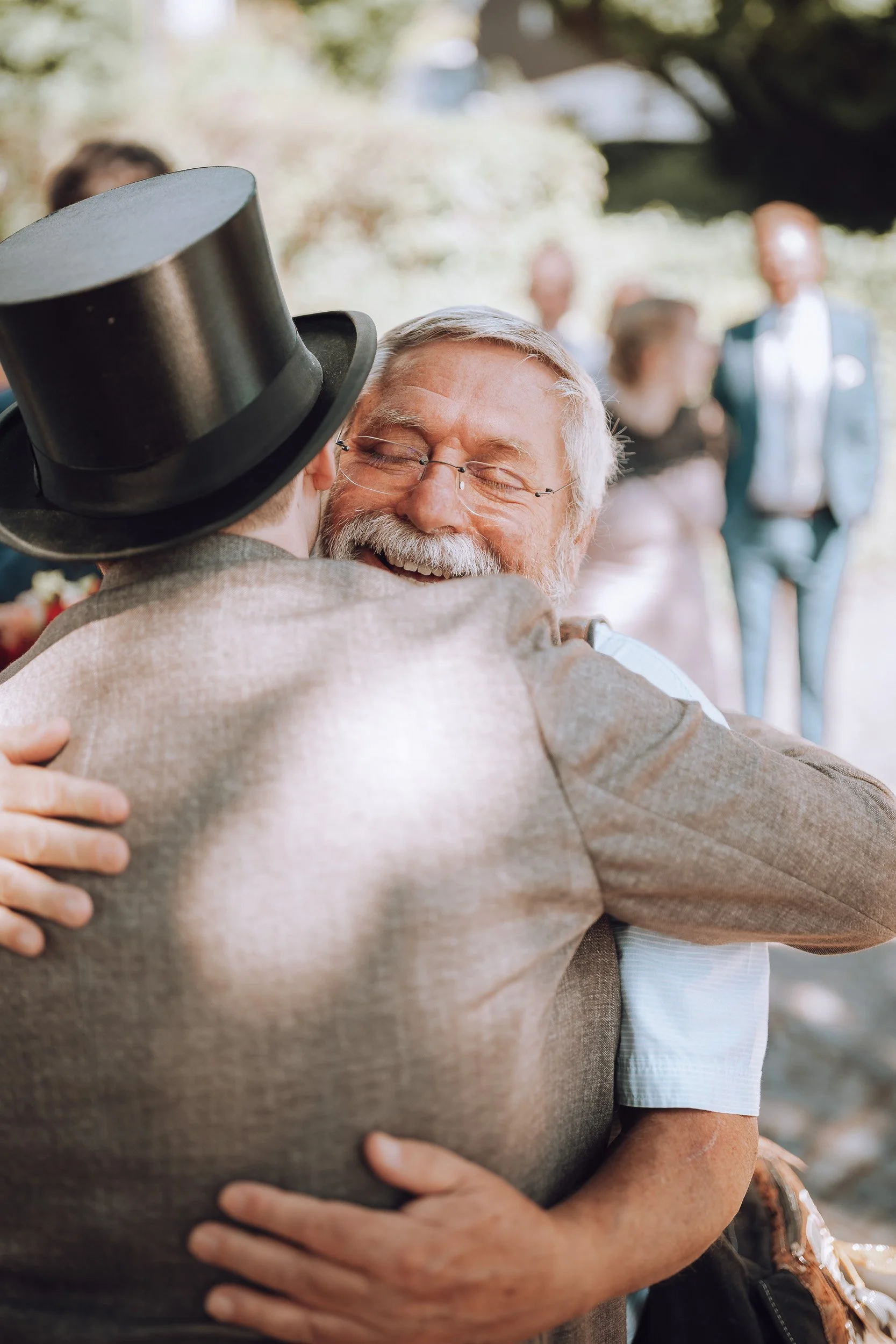Zwei Männer umarmen sich herzlich bei einer outdoor Feier, einer trägt einen schwarzen Zylinderhut und mit einem Lächeln