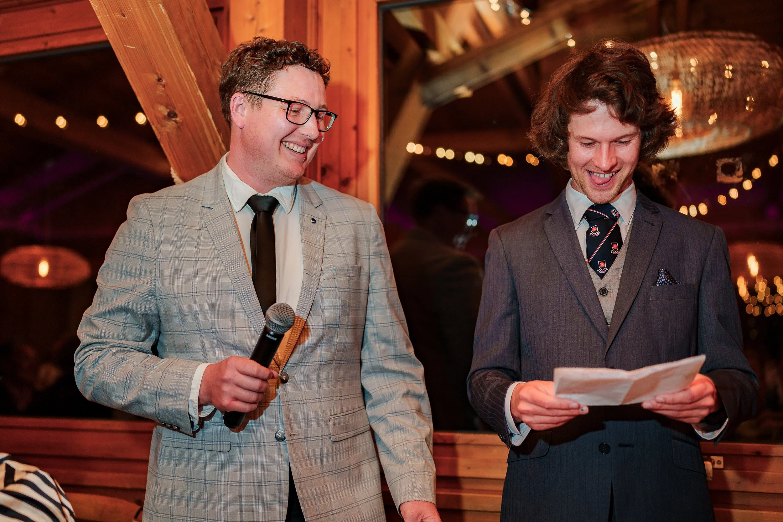 Zwei Männer in Anzügen bei einer Feier, einer hält ein Mikrofon, beide lachen und schauen auf ein Stück Papier, im Hintergrund Lichter und eine Holzwand