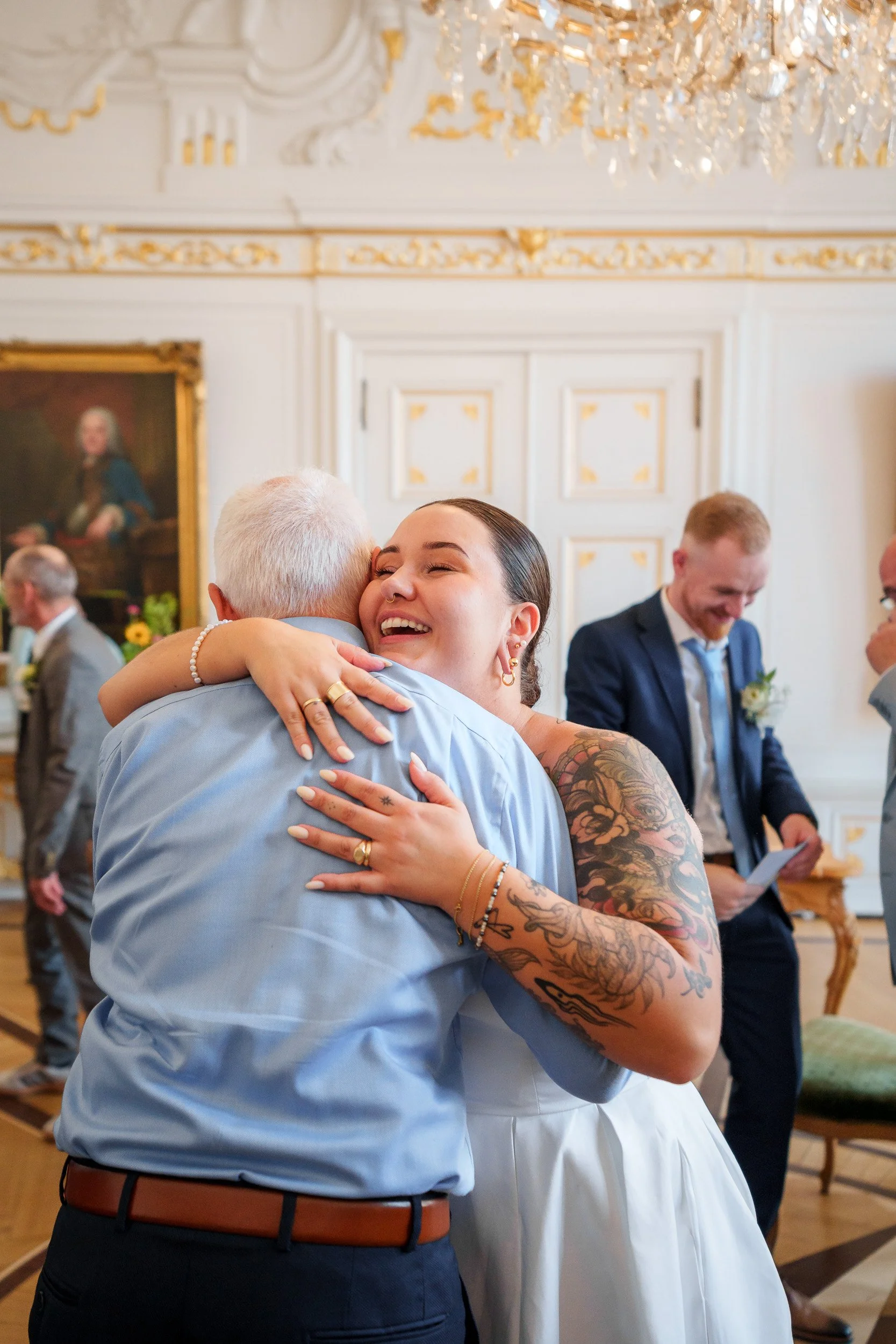 Eine glückliche junge Frau mit Tattoos umarmt einen älteren Mann bei einer Hochzeit im eleganten, historischen Saal mit weißer und goldener Dekoration.