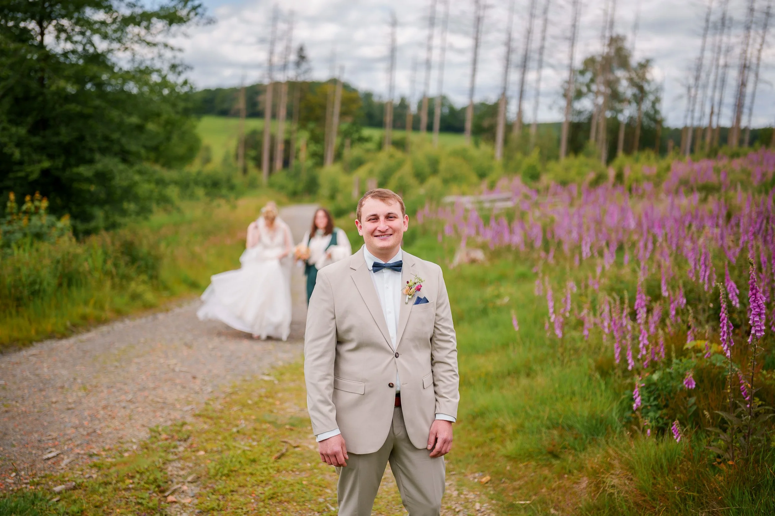 Ein Mann in einem beige Anzug, mit Fliege, steht vor einer ländlichen Kulisse mit Blumen, im Hintergrund eine Braut und eine weitere Person, alle bei einer Hochzeitsaufnahme im Freien.