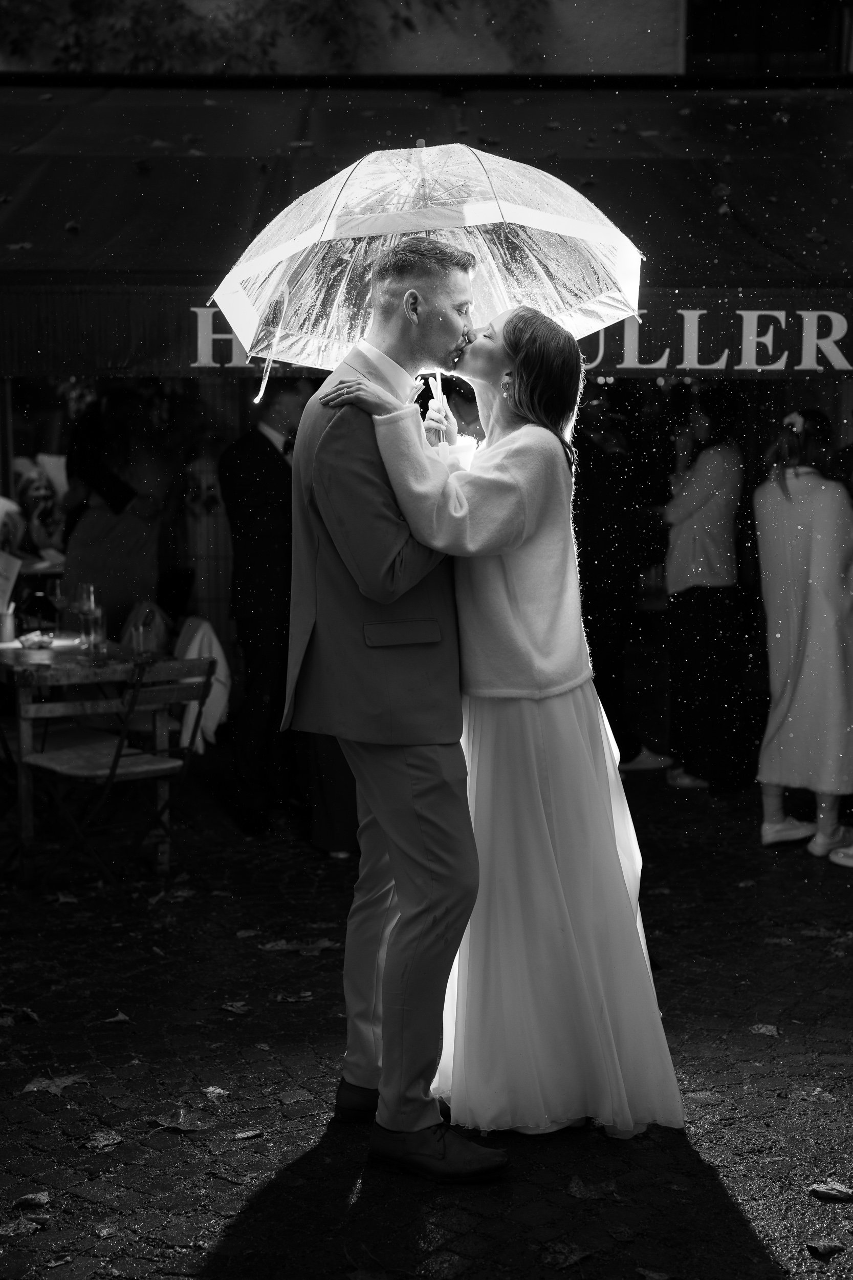 Ein Paar küsst sich unter einem transparenten Regenschirm bei Regen während einer Hochzeit oder Feier, im Hintergrund sind Gäste und ein Firmenlogo sichtbar.