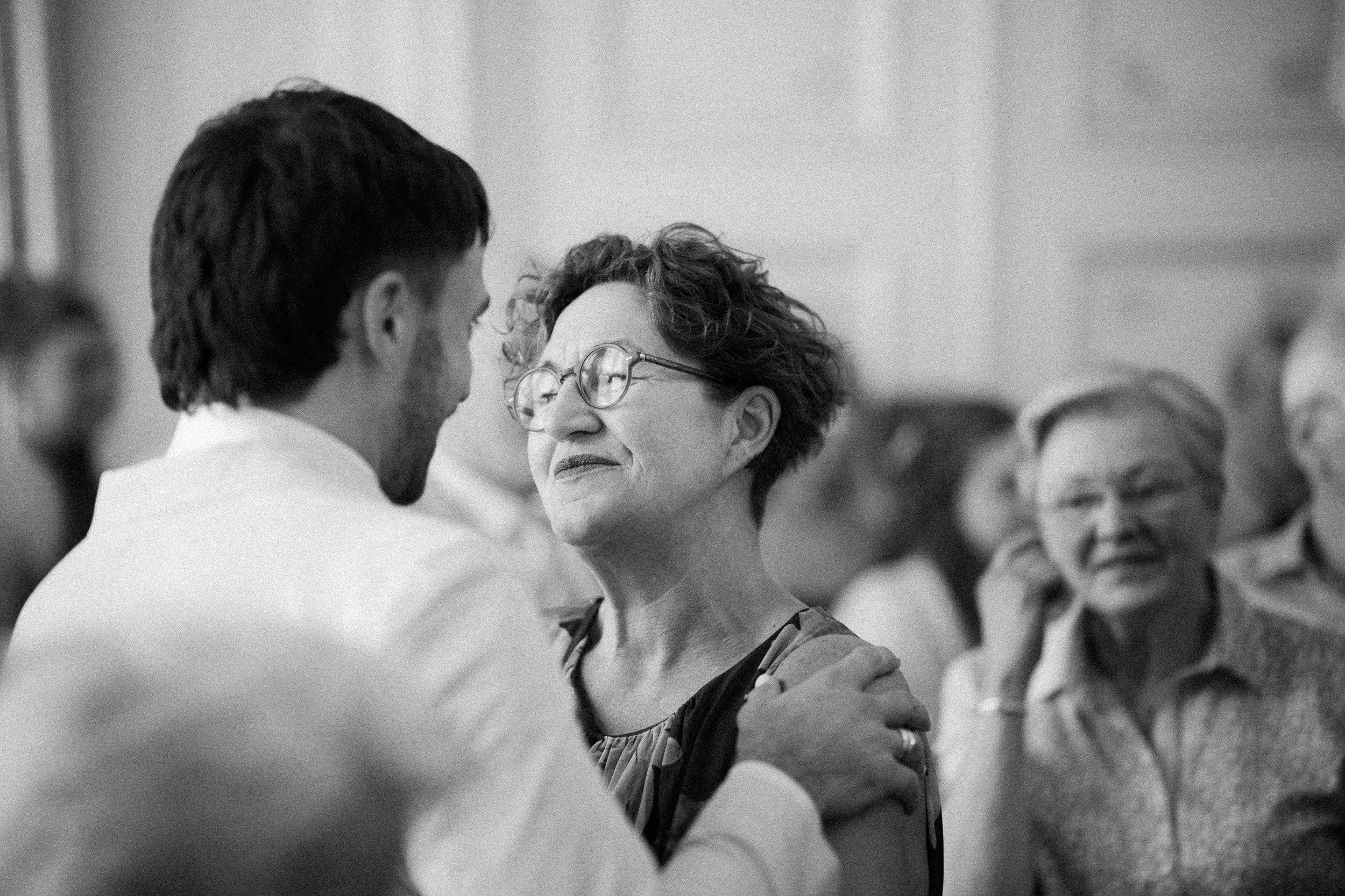 Ein Mann hält eine ältere Frau im Arm, die in einer Andacht oder einem Tanz erleichert und glücklich aussieht, während andere Menschen im Hintergrund zusehen.