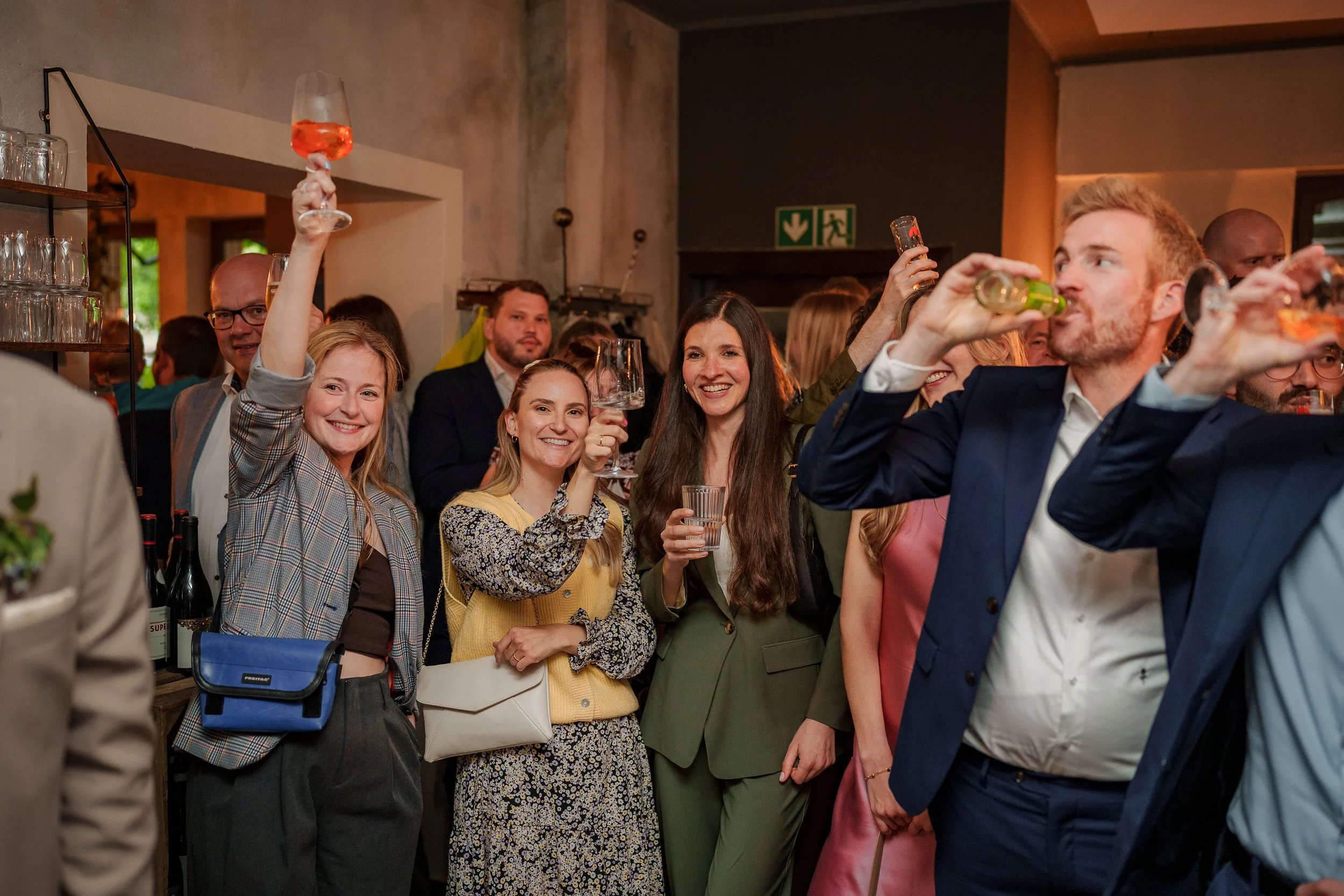Menschen bei einer Feier, die Getränke in Gläsern und Flaschen trinken, lachen und anstoßen, in einem festlichen Umfeld mit bartierten und modernen Kleidung.