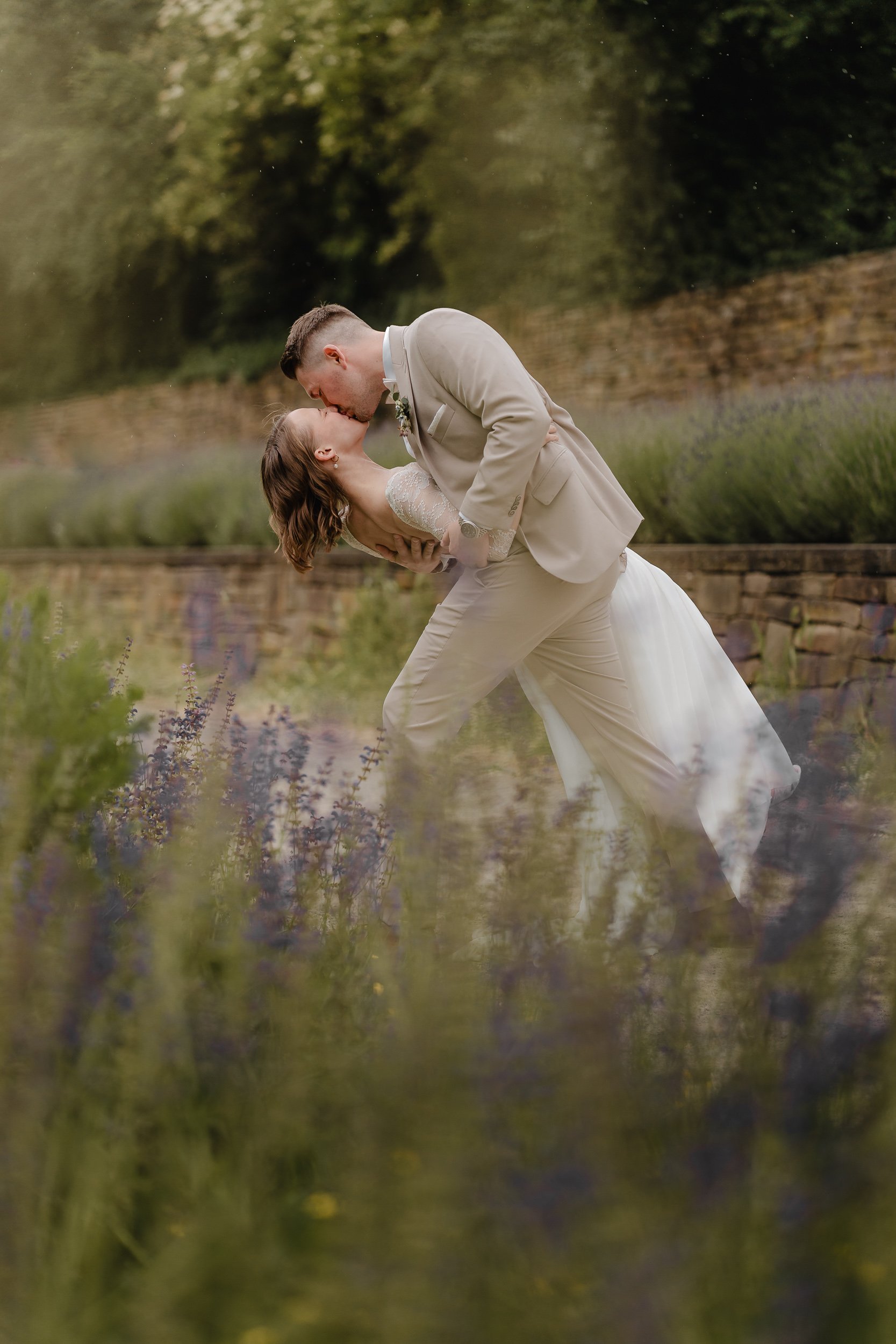 Ein Brautpaar teilt einen Kuss im Freien an einem Bach, umgeben von Natur mit Büschen und Bäumen.